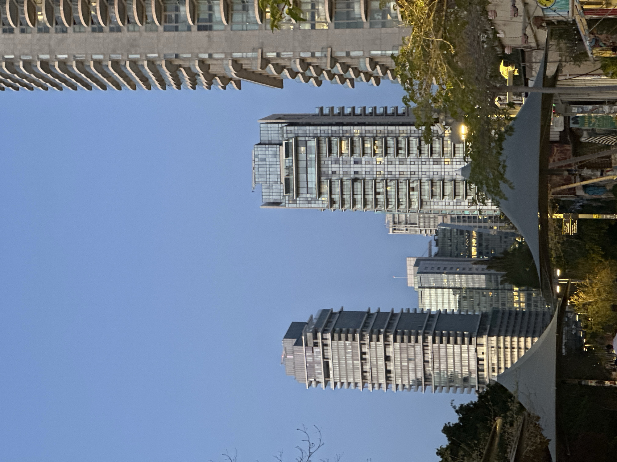 This media file captures a modern urban landscape at dusk or early evening, with tall buildings silhouetted against a clear, light blue sky. The original image is oriented horizontally, with the buildings appearing vertically when rotated correctly.
In the prominent left portion of the frame (when rotated), a distinctive high-rise building features a facade with numerous, uniformly wavy or scalloped balconies, giving it a textured and rhythmic appearance. To its right, another tall building with a sleek glass and concrete exterior reflects the ambient light, and many of its windows are illuminated, suggesting interior activity. Further back and to the right, a third contemporary skyscraper with a more complex, segmented facade also displays illuminated windows. Several other modern high-rise buildings are visible in the background, contributing to the urban density.
The right side of the image (when rotated) reveals ground-level details. A large, light-colored fabric canopy or awning occupies the lower right, partially obscuring the view. Around and behind this structure, lush green trees with visible branches add natural elements to the scene. Several streetlights are illuminated, casting warm light on the area. The setting appears to be a public space or boulevard within a bustling city.
The sky is a uniform, clear light blue, indicative of fair weather as the day transitions to night. No specific activities or individuals are clearly discernible. While there are some signs visible in the background, any text on them is not legible. The scene depicts a contemporary city, consistent with locations like Tel Aviv, Israel, known for its modern architecture and vibrant urban environment.