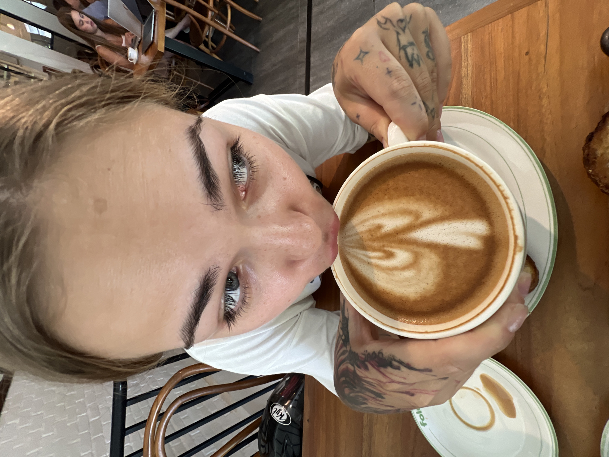 A person is drinking a cappuccino from a white cup with a green-rimmed saucer. The cappuccino features latte art, and the person is holding the cup with both hands. The person has light skin, dark eyebrows, and blue eyes, wearing a white shirt. The person has tattoos on both hands and arms. A table is visible under the cup and saucer. In the background, another person is sitting at a table with a laptop. The location is likely a cafe in North Kuta, Indonesia, although no specific landmarks are visible.