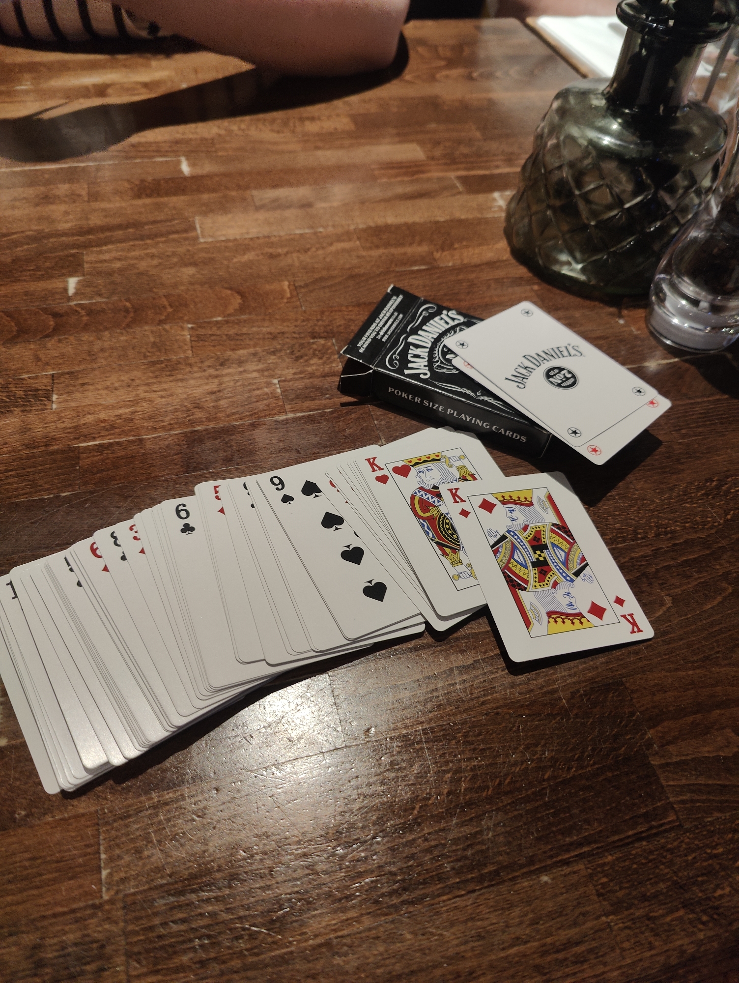 On a wooden surface, possibly a table, a deck of playing cards is scattered, along with its box and a decorative glass bottle. The playing cards, with a white background and red/black suits, are splayed out, revealing various values like an Ace, three, six, nine, and several Kings. The box for the playing cards is black and white, featuring the Jack Daniel's logo and text indicating they are poker-sized. A decorative glass bottle with a dark green tint and a geometric pattern sits nearby.
