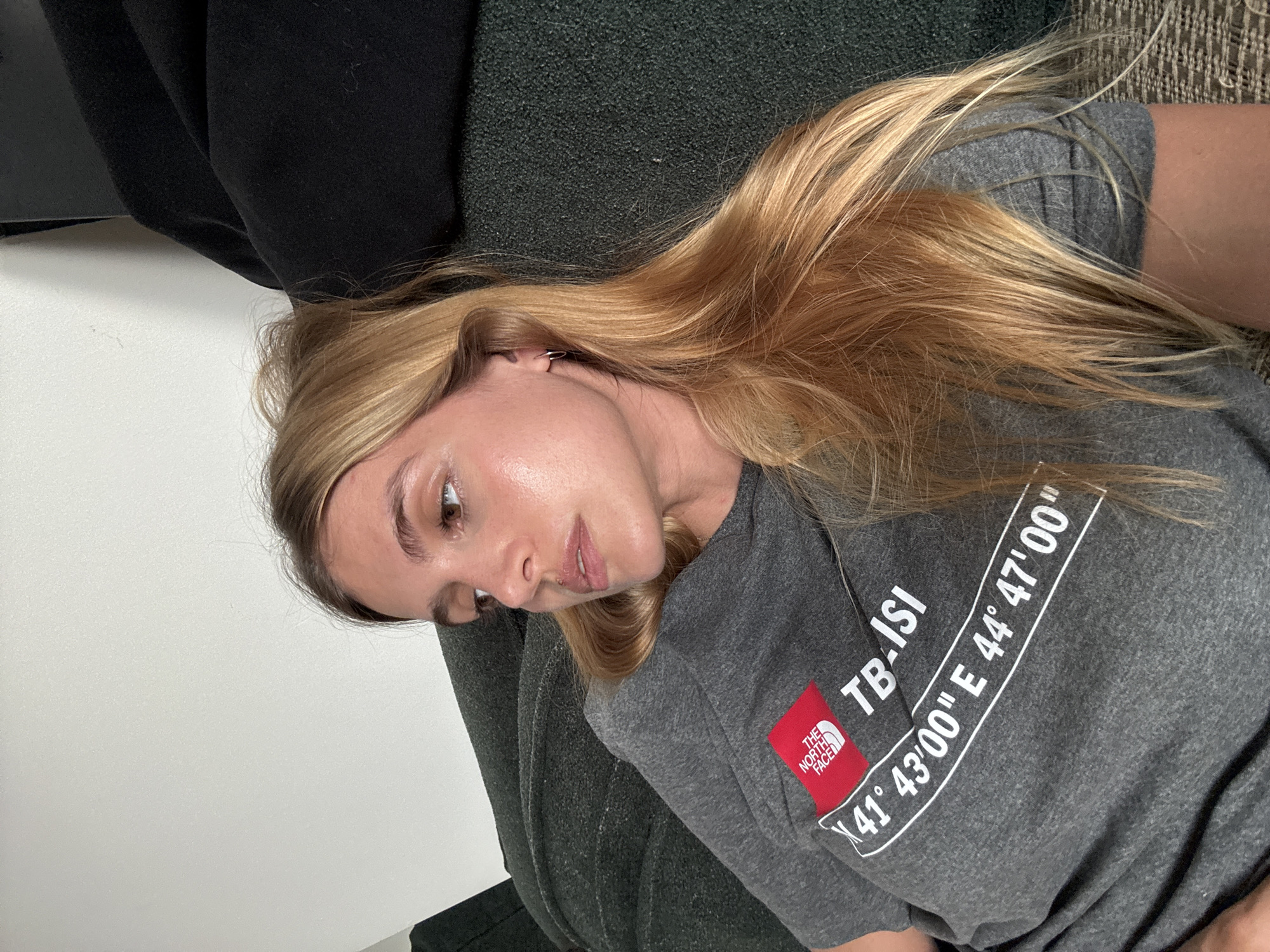 A woman is lying down, facing slightly to the left. She has light skin and long, blonde hair that is tousled. She's wearing a grey t-shirt with a red North Face logo, and text that reads "TB-ISI" and the coordinates "41° 43'00" E 44° 47'00"." Her head is resting on a dark green, textured surface, possibly a cushion or couch. There's a white wall visible in the background to the left. The coordinates on the shirt correspond to Tbilisi, Georgia.
