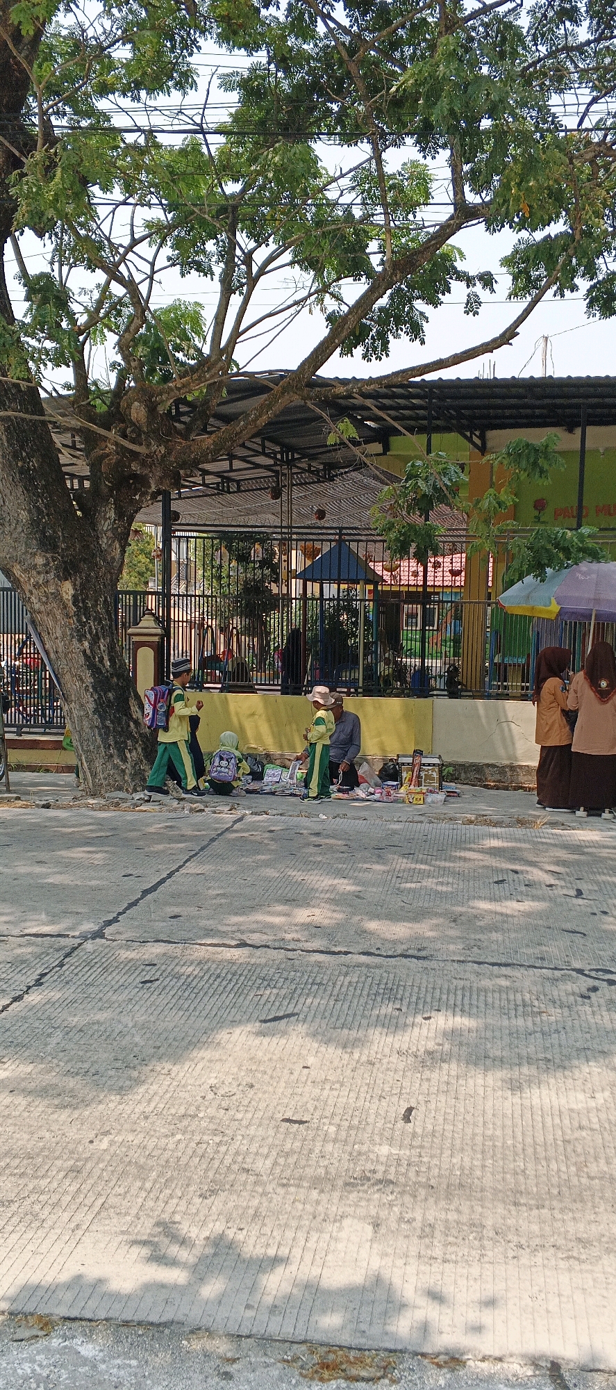 In the city of Karangwungu, Indonesia, a street scene unfolds under a large tree with dappled sunlight casting shadows on the concrete pavement. A vendor sits on the ground next to a collection of merchandise, including books and various items displayed on the ground. Two children, dressed in school uniforms with green and yellow colors and backpacks, are present. One child stands and appears to be interacting with the vendor, while another child is crouched down looking at the merchandise. To the right, two women in hijab, one in an orange outfit and the other in a beige outfit, are standing near a striped umbrella. In the background, a fence encloses a schoolyard with playground equipment. A large tree trunk is prominently visible on the left side of the image.