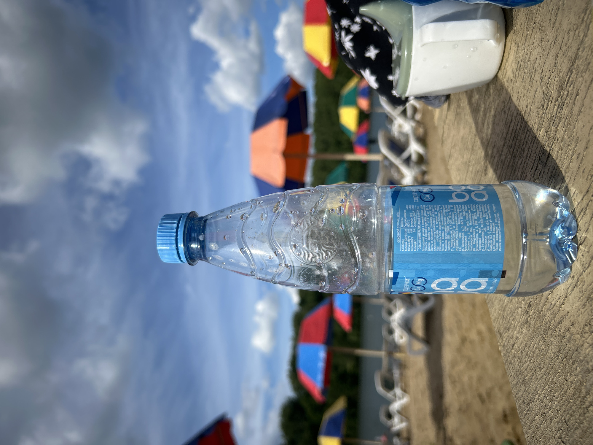 In the foreground, a clear plastic bottle of water with a blue cap stands on a weathered wooden surface. The bottle has a blue label with white text and the logo "BonAqua". Water droplets are visible on the exterior of the bottle. To the right of the bottle, a white sippy cup with a green lid is partially visible, also with water droplets. In the background, a sandy beach is lined with lounge chairs and colorful beach umbrellas in red, blue, orange, and yellow. The water appears calm, and trees and a hill are visible on the opposite bank of the body of water. The sky is partly cloudy with blue sky visible between the clouds. The scene depicts a relaxing day at a beach or lakeside, possibly in the city of Yubileynyy, Russia, given the labeling on the bottle. There are no people clearly visible in the image.