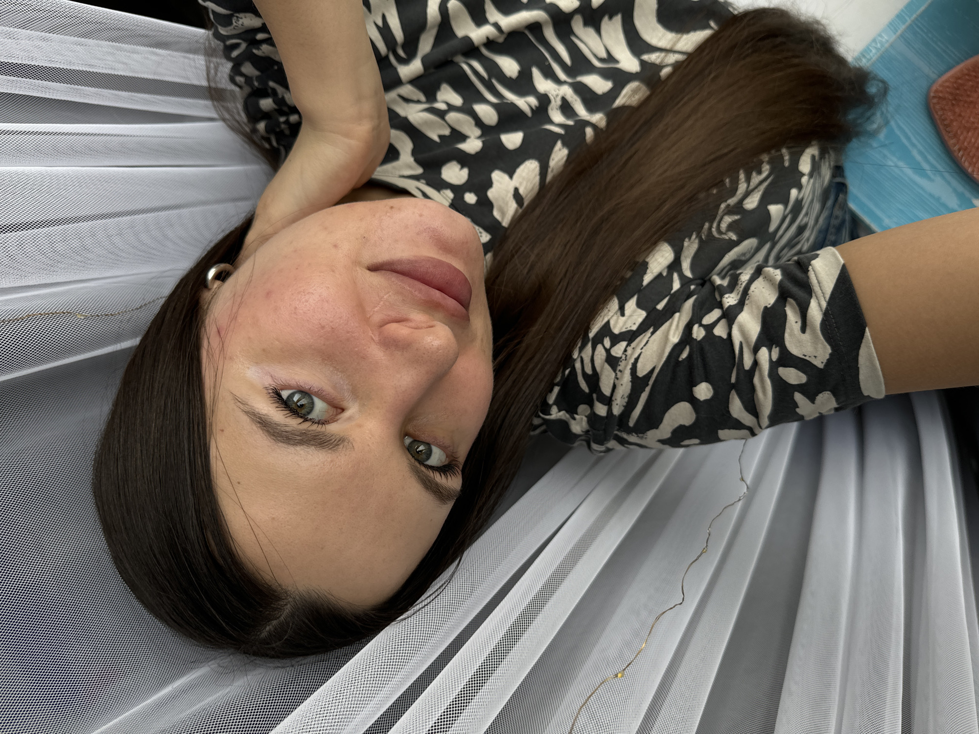 A woman with long, straight dark brown hair and greenish-hazel eyes is depicted in a close-up, eye-level shot, lying down and looking directly into the camera. She has a neutral expression and visible skin texture on her cheeks. She is wearing a black and off-white/beige patterned top, possibly an abstract or animal print, and a simple silver hoop earring in her left ear. Her left arm is bent, with her hand positioned near her head.
She is lying on or under a white, sheer, finely-meshed fabric, resembling tulle or netting, which is draped in soft folds around her. A delicate string of tiny, warm-toned fairy lights is subtly intertwined within this white fabric, visible on the right side of the frame. In the upper right background, parts of a blue patterned surface and a reddish brush can be seen, indicating an indoor setting. The image is brightly and evenly lit. There is no visible text.
Location context: Unknown, Unknown