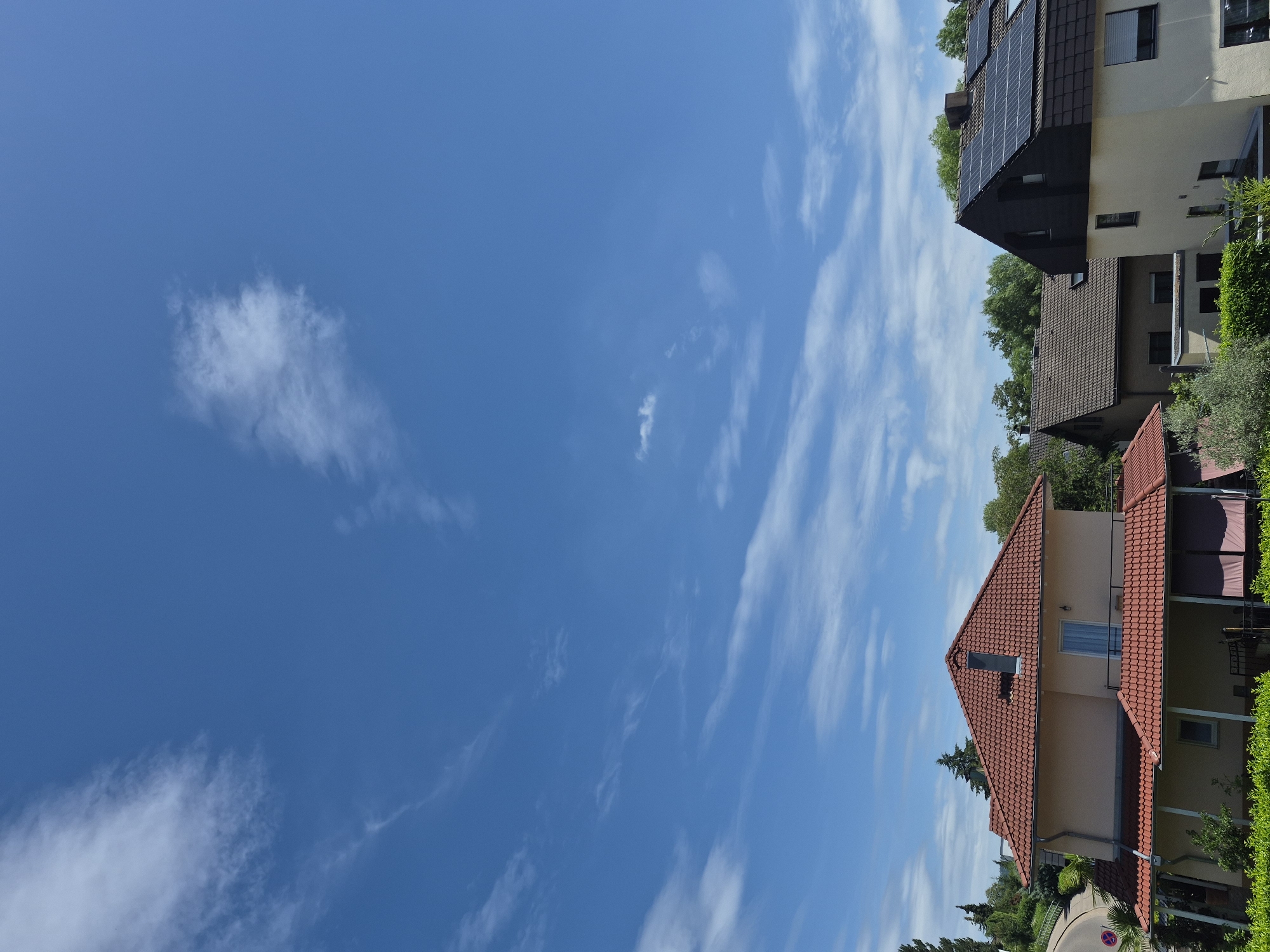 The image shows a residential scene with several houses under a bright blue sky with wispy white clouds. The houses have red tile roofs and are surrounded by green trees and foliage. One house in the foreground has a light-colored facade and a covered patio area. Another house behind it has a darker roof and solar panels on its roof. The image appears to be taken on a clear, sunny day, likely in the afternoon given the brightness of the sky. The perspective is from a slightly elevated position looking down on the houses. There are no people visible in the image. There is a small red and white circular sign with a red slash on it visible near the bottom edge, which is likely a traffic or parking sign. The overall mood is peaceful and tranquil.