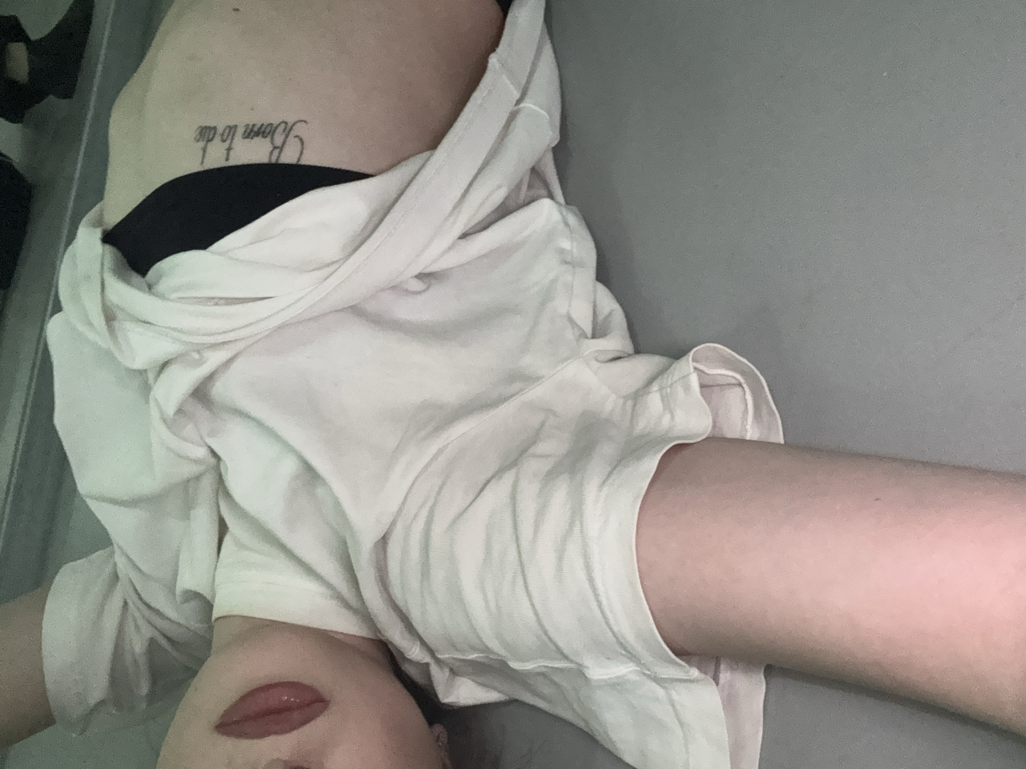 A medium shot of a person lying down. The person has a tattoo on their side that says "Born to die". The person is wearing a white shirt and black underwear. The lips are prominent. The background is a gray wall. There is no evidence to indicate this image was taken in Sofia, Bulgaria.