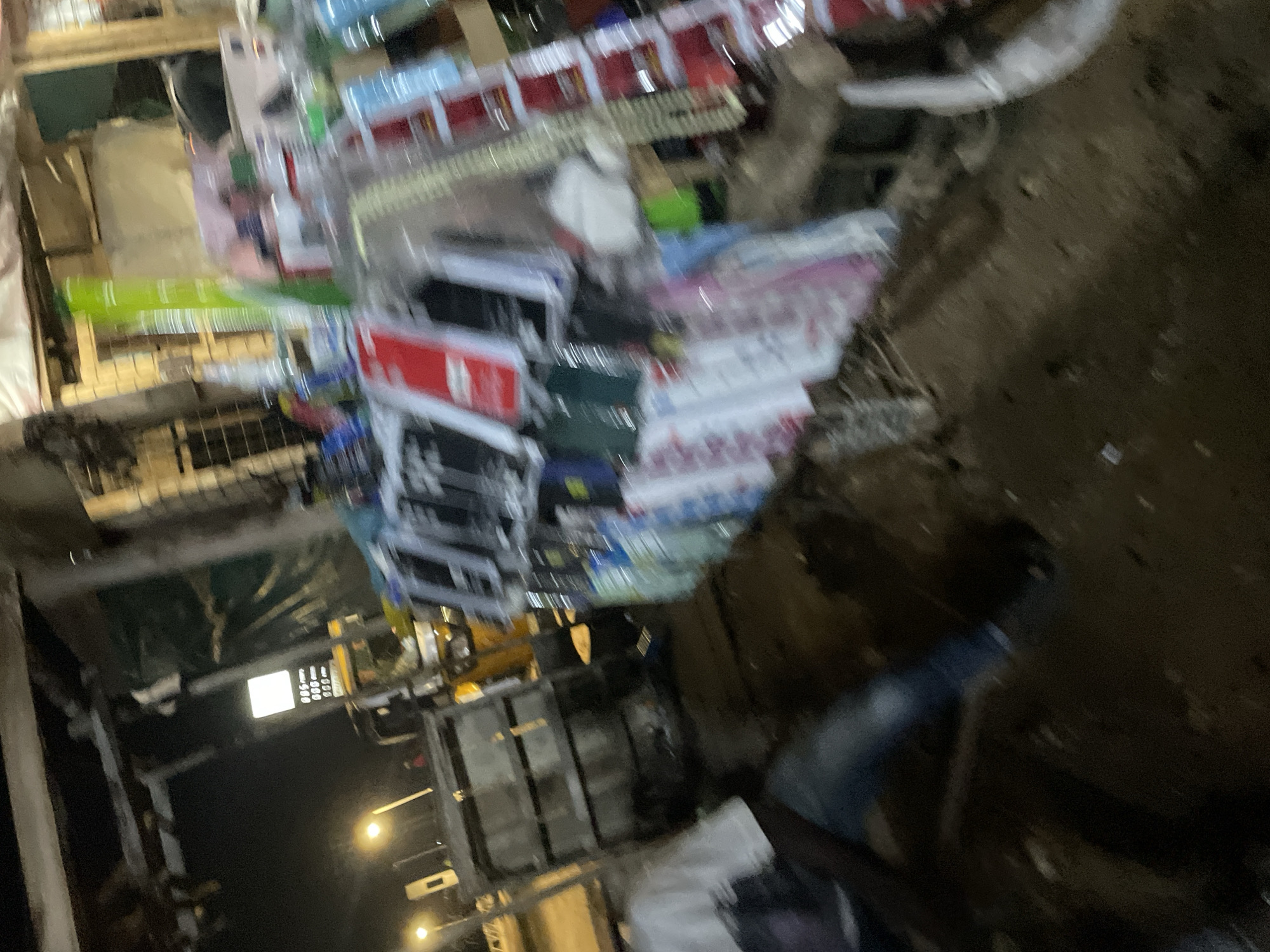 The image depicts an outdoor scene at night, likely a market stall or roadside vendor in Lokoja, Nigeria. In the foreground, a person wearing a white t-shirt and blue jeans is partially visible, seated or crouching. Behind them, a collection of items is displayed, appearing to be mobile phone accessories such as cases, and possibly other small electronics. These items are arranged on tables or shelves, some of which are made of wood. The background shows a structure that resembles a stall or shop with a roof and wooden supports. A brightly lit sign with numbers, possibly indicating prices or fuel costs, is visible in the midground. Streetlights are also present, illuminating parts of the scene. The ground is dark and appears to be unpaved, with some debris scattered around. The overall impression is one of a typical evening market setup in an urban or semi-urban environment.