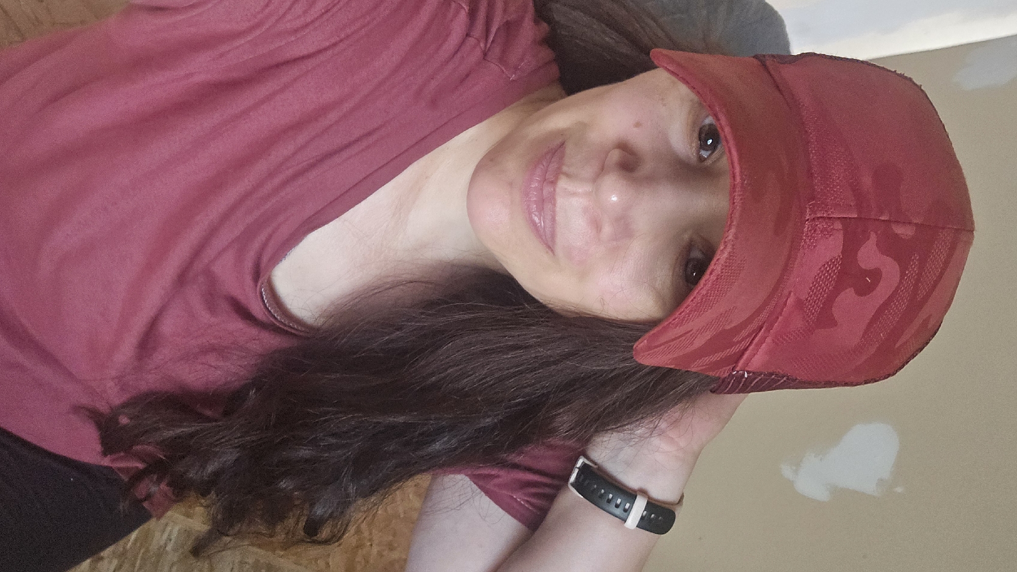 A woman with long, dark hair is wearing a red, camouflage-patterned baseball cap and a maroon t-shirt. She is resting her right hand on the back of her head, and a black and white fitness tracker is visible on her left wrist. The background appears to be an indoor environment with a beige wall that has some patches of white repair work. The woman is looking directly at the camera with a slight smile.