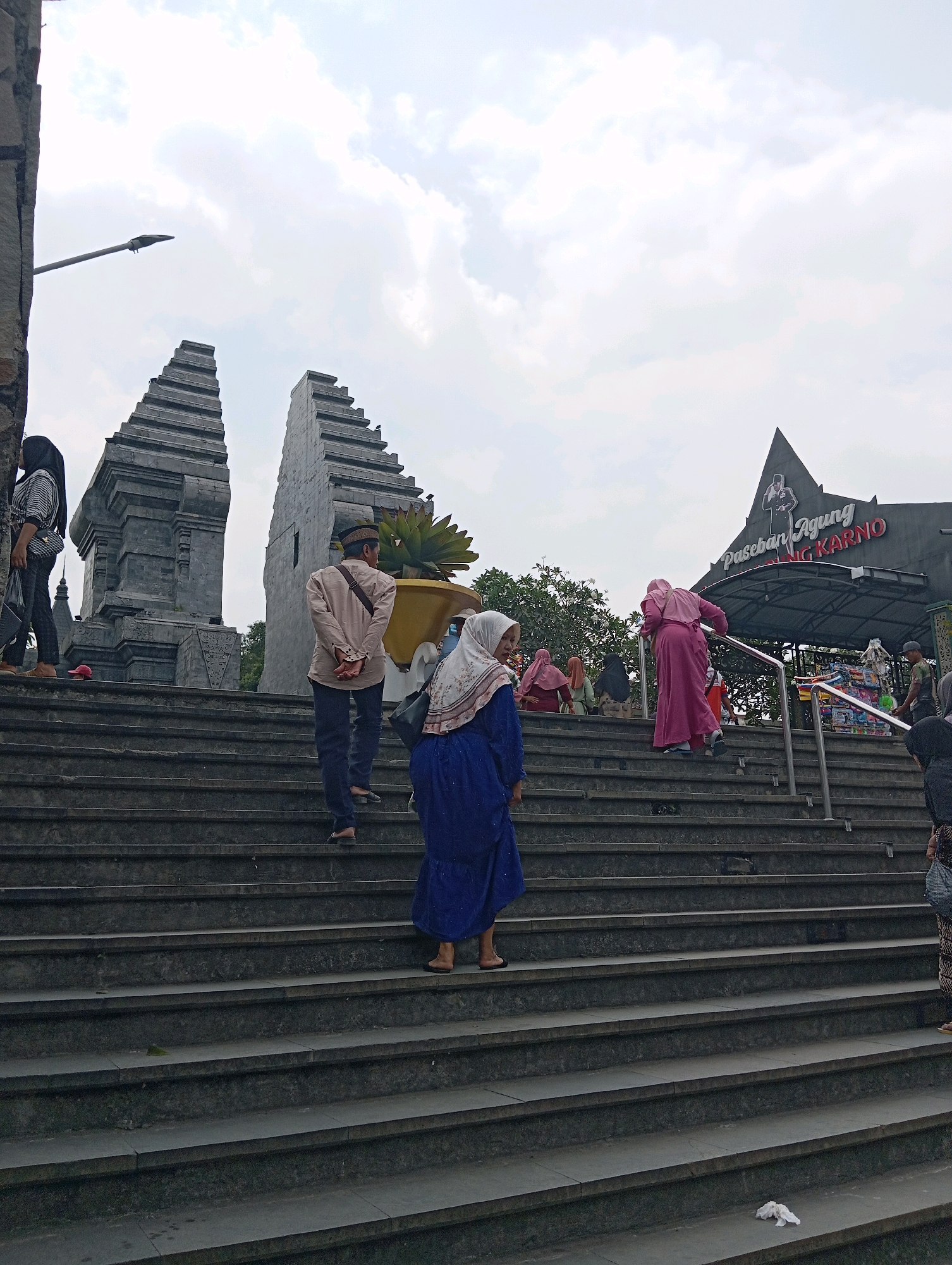 In Blitar, Indonesia, a wide outdoor staircase leads up to a building with a prominent sign reading "Paseban Agung Ir. Soekarno." The staircase is made of dark gray stone, with a slight sheen suggesting it may be wet or polished. Several people are ascending the stairs. On the left side of the stairs, a woman in a striped hijab and a black and white striped shirt walks upwards. Further up, a man in a light brown long-sleeved shirt and dark pants, wearing a traditional cap, also walks up. In the center of the frame, a woman in a vibrant blue dress and a light-colored hijab walks up the stairs. Another woman in a pink hijab and pink dress is climbing the stairs on the right side. Behind the stairs, there are two large, tiered stone structures that resemble ancient gateways. The sky is overcast with visible clouds. To the right, a black awning extends from the building, and a large silhouette of a man, presumably Ir. Soekarno, is visible against the sky. Shops are set up under the awning, with colorful goods displayed. The overall scene depicts a popular tourist or cultural site.