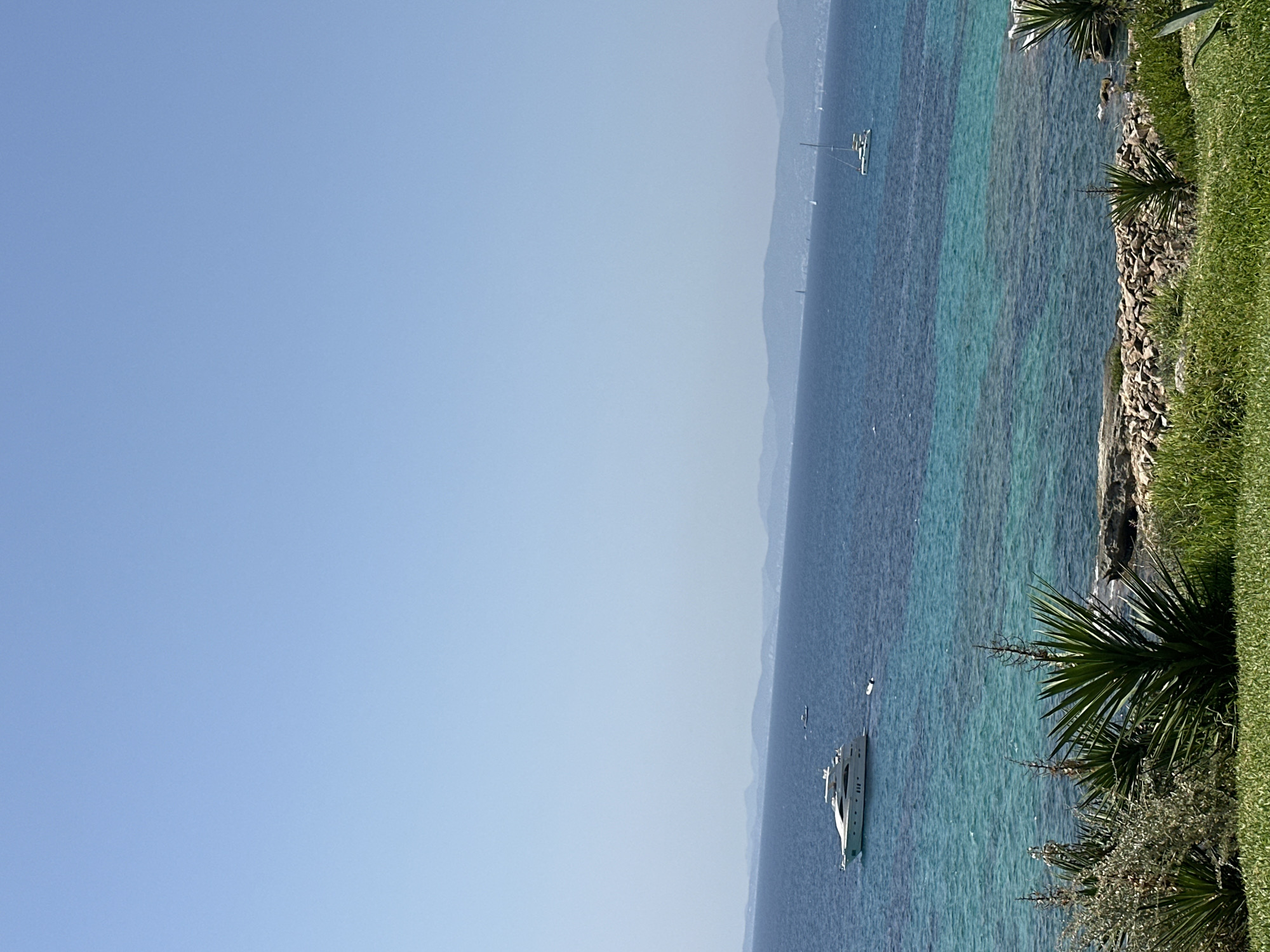 This is a photograph taken from a high vantage point overlooking a coastal scene. The image is dominated by a clear, pale blue sky which occupies the upper two-thirds of the frame. Below the sky, a vast expanse of deep blue ocean stretches across the horizon. In the middle ground, a yacht is anchored in the water, appearing white and sleek. Several smaller boats are also visible, some closer to the shore and others further out at sea.
The water transitions from deep blue to a vibrant turquoise closer to the shore, indicating shallow, clear water. The coastline is lined with rocky areas and patches of green grass. In the foreground, lush green grass covers the lower part of the frame, with some spiky, green plants growing amongst the rocks and grass. In the distance, a hazy mountain range can be seen along the horizon.
The location appears to be a coastal area in the municipality of Llucmajor, Spain, likely on the island of Mallorca. The scene depicts a tranquil day at sea with several vessels enjoying the water. There are no people clearly visible in the image.