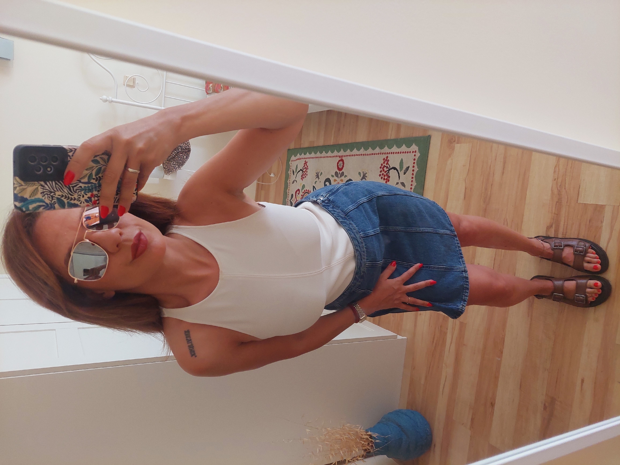 A woman is taking a mirror selfie in what appears to be a bedroom. She is wearing a white tank top, a denim skirt, and brown sandals. She has red nail polish and is wearing sunglasses and a ring. A phone with a floral case is held in her right hand. A tattoo is visible on her left shoulder. In the background, there is a white wardrobe, a bed with a white headboard, and a rug with a floral pattern. A blue vase with dried plants sits on the floor to the left. The flooring is light-colored wood. The room is well-lit, likely by natural light. There is no indication of the city of Sozopol, Bulgaria, in the image.
