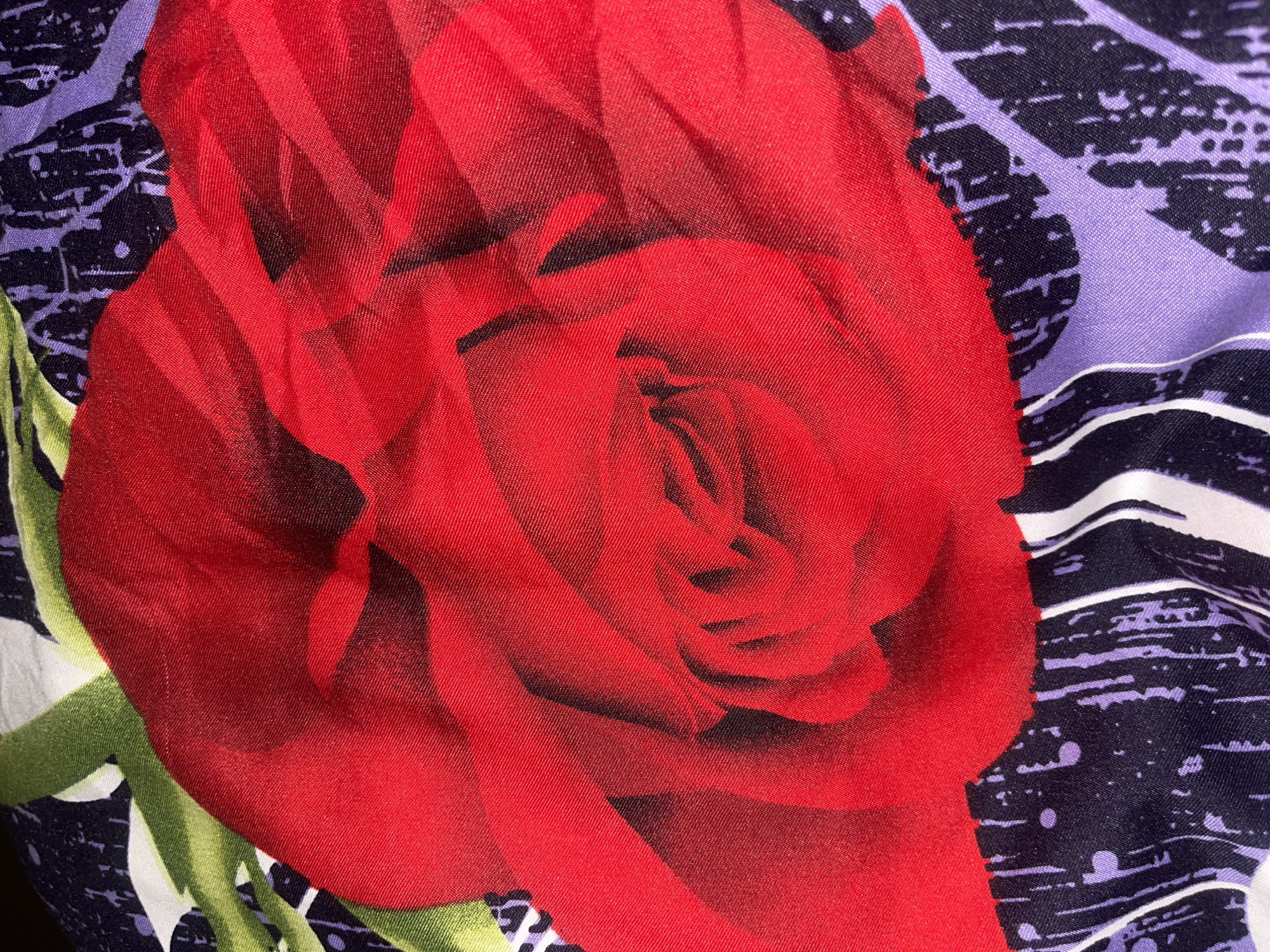 The image is a close-up, overhead view of a fabric with a floral print. A large, red rose dominates the center of the frame, its petals creased and layered. The background fabric features a pattern of abstract, distressed shapes in black and purple, interspersed with vertical white stripes. Green leaves are visible around the edges of the rose. The fabric appears to be draped or folded, creating shadows and highlights that emphasize its texture. The scene is not identifiable as being located in Minsk, Belarus, or any other specific geographic location.