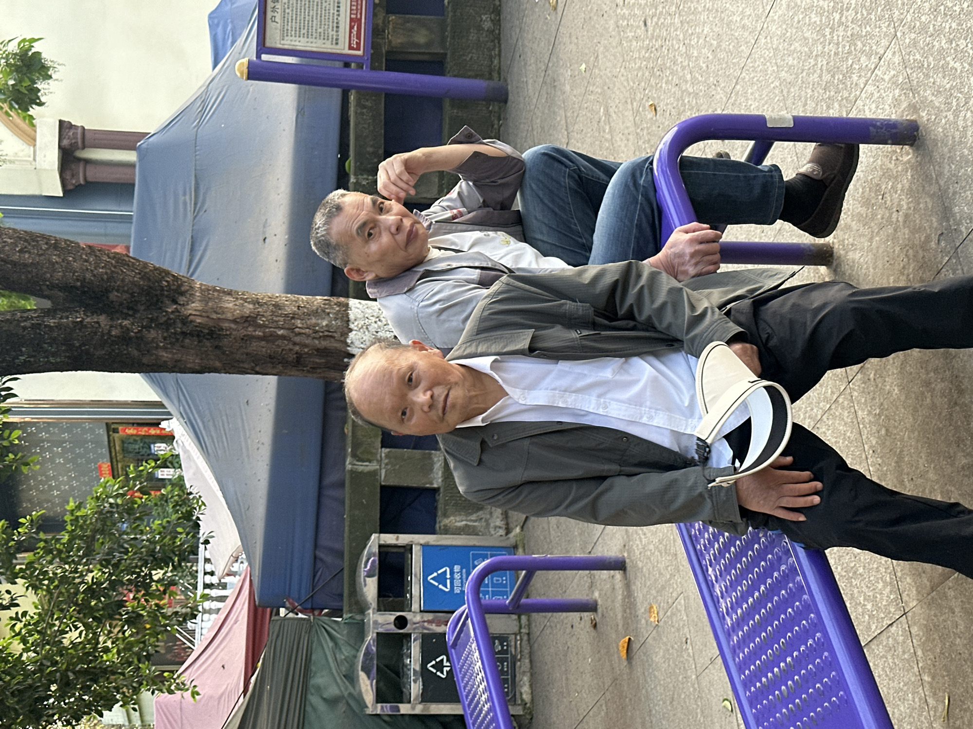 Two older Asian men are seated on a vibrant purple metal outdoor bench in a public space in Jiangjin, China, both looking directly towards the camera.
The man in the foreground wears a white collared shirt, a dark olive green jacket, and black pants. He holds a white and black hat in his lap with his left hand, which also rests on his thigh. His expression is neutral. Behind him, the second man, with grey hair, is dressed in a light-colored t-shirt with a yellow graphic, an open light grey jacket, and blue jeans. His left arm is bent, with his hand resting on his left knee, and he also maintains a neutral gaze.
To the left, a large tree trunk, marked with a white band, partially frames the scene. In the middle ground behind the men, various tarpaulins or covers in shades of blue, red, and green are visible, suggesting market stalls or temporary structures. Further back, architectural elements like columns can be seen. The ground is paved concrete, scattered with a few fallen leaves.
Adjacent to the bench and behind the foreground man, two public trash bins are visible: a grey one and a blue recycling bin. The blue bin is clearly marked with the recycling symbol and the Chinese characters "可回收物" (kě huíshōu wù), meaning "recyclable materials." A partially visible red and white sign in the upper right corner includes the Chinese characters "户内" (hùnèi), meaning "indoor" or "household." The scene is lit by bright daytime light, indicating a clear day.