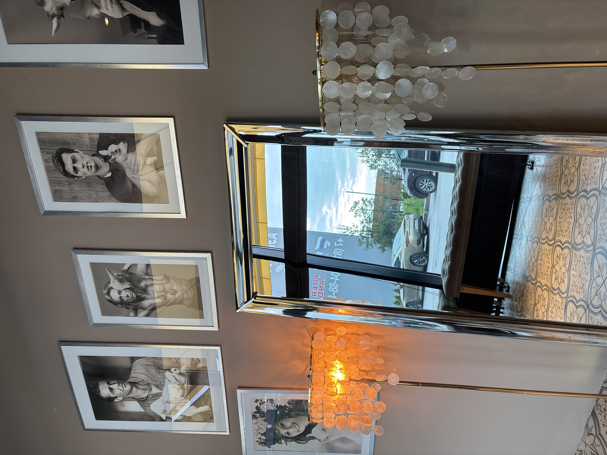A photograph taken indoors shows a beige wall adorned with five framed black and white photographs, and a large silver-framed mirror reflecting a street scene.
The photographs feature various subjects, including what appears to be a man with a dog, a woman, and another woman with a baby. The frames are a uniform silver color.
To the right, a long, rectangular mirror reflects an external view, including part of a building with lettering in both English and Cyrillic script. The reflection also shows part of a street with cars and greenery. The lettering visible on the building in the mirror includes part of a phone number that starts with "+7(8)."
A decorative lighting fixture composed of cascading translucent discs is affixed near the mirror's edge on both the top and bottom.
Based on the Cyrillic lettering and the "+7" phone code, the location is likely in Russia. Considering the street scene and architecture, the city might be Saint Petersburg.