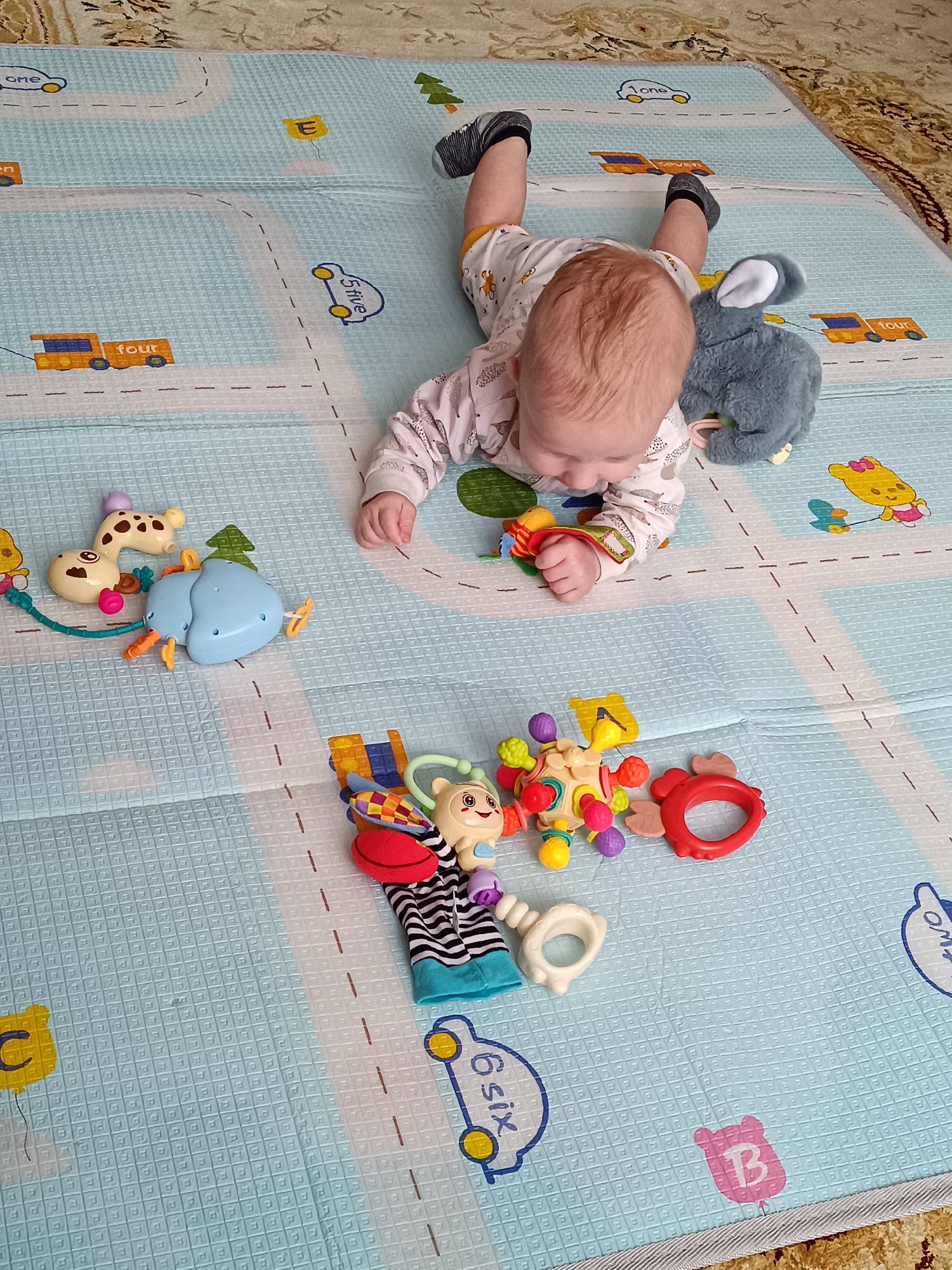 A baby wearing black socks and a patterned onesie is lying on their stomach on a light blue playmat with white roads and cartoon cars. The baby has light skin and reddish hair and is reaching for a toy in front of them. Several other toys are scattered around the baby on the playmat, including a blue elephant, a giraffe, a stuffed gray bunny, and various plastic rattle-like toys. The playmat is on a carpet with a tan and brown pattern. There is no indication in the image that suggests that it was taken in the city of Tobolsk, Russia.