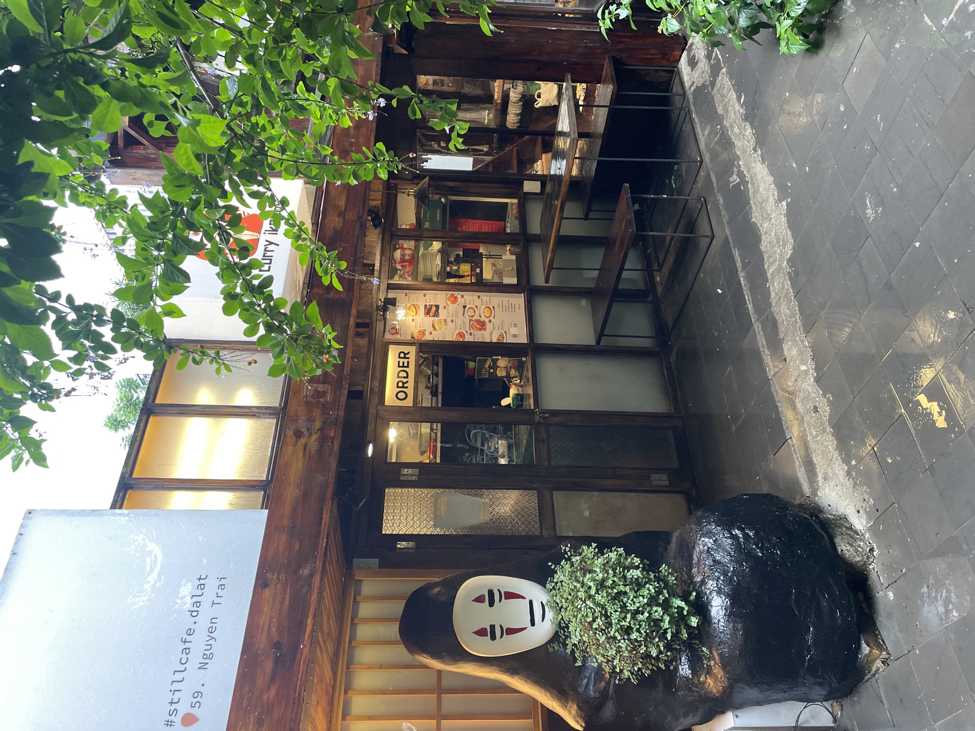 The image depicts the exterior of a café in Lâm Viên - Đà Lạt, Vietnam. The café's name, "#stillcafe.dalat", and address, "59 . Nguyen Trai", are displayed on a sign. The building features a wooden facade with glass windows and an "ORDER" sign. A decorative sculpture resembling a face mask and covered with greenery is positioned near the entrance. The sidewalk is tiled and wet. The photograph is taken from a low angle, with foliage visible in the upper left corner.
