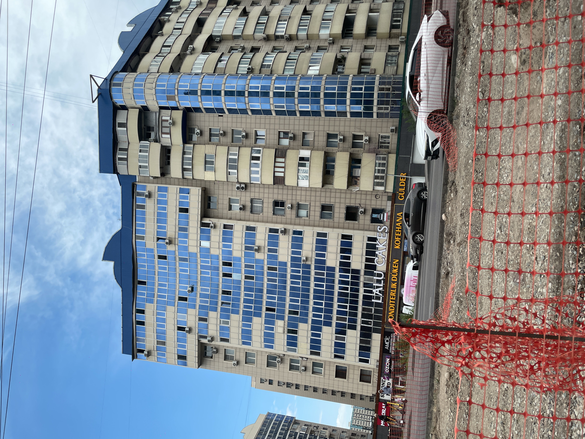 The image shows a street view in Almaty, Kazakhstan, with a tall, multi-story building featuring a beige facade and numerous blue-tinted windows. The building has a distinctive blue, curved roofline and several balconies. In the foreground, a red mesh safety fence is visible, with construction debris and gravel behind it. A street runs in front of the building, with several cars on it. One car, a white sedan, is prominent in the right side of the frame, partially obscured by the fence. Signs on the building's ground floor include "LALU CAKES," "ONDITERLIK DÜKEN," and "KOFEHANA." The sky is partly cloudy, and power lines are visible crossing the upper portion of the image.