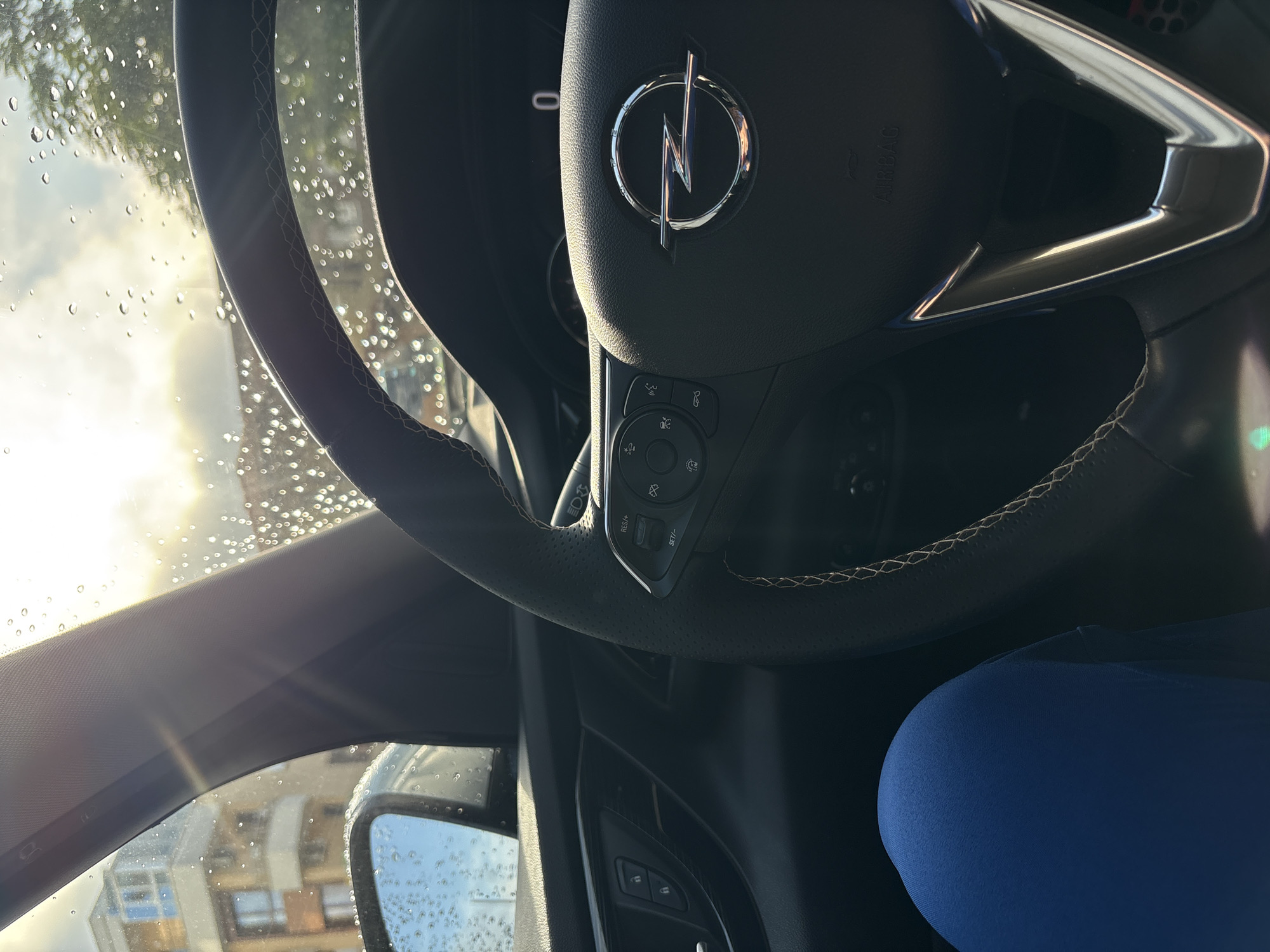 A low-angle shot captures the interior of a car, specifically focusing on the steering wheel and the view through the windshield and driver's side window. The steering wheel, dark grey with light stitching, prominently displays the chrome Opel logo with its distinctive lightning bolt symbol in the center. To the left of the logo, the word "AIRBAG" is visible. The right spoke of the steering wheel features various control buttons with icons for phone calls, volume adjustment, and cruise control (including "RES/+" and "SET/-").
Through the windshield, bright sunlight streams in, creating lens flares and illuminating numerous water droplets adhering to the glass, suggesting recent rain or dew. Silhouetted against the bright sky are tree branches. On the driver's side, outside the car, part of a building with windows and a side mirror also covered in water droplets are visible. A blue-clad leg or lap is seen in the bottom right corner of the frame. The overall setting appears to be a daytime scene in Plovdiv, Bulgaria, with the vehicle either parked or stationary.