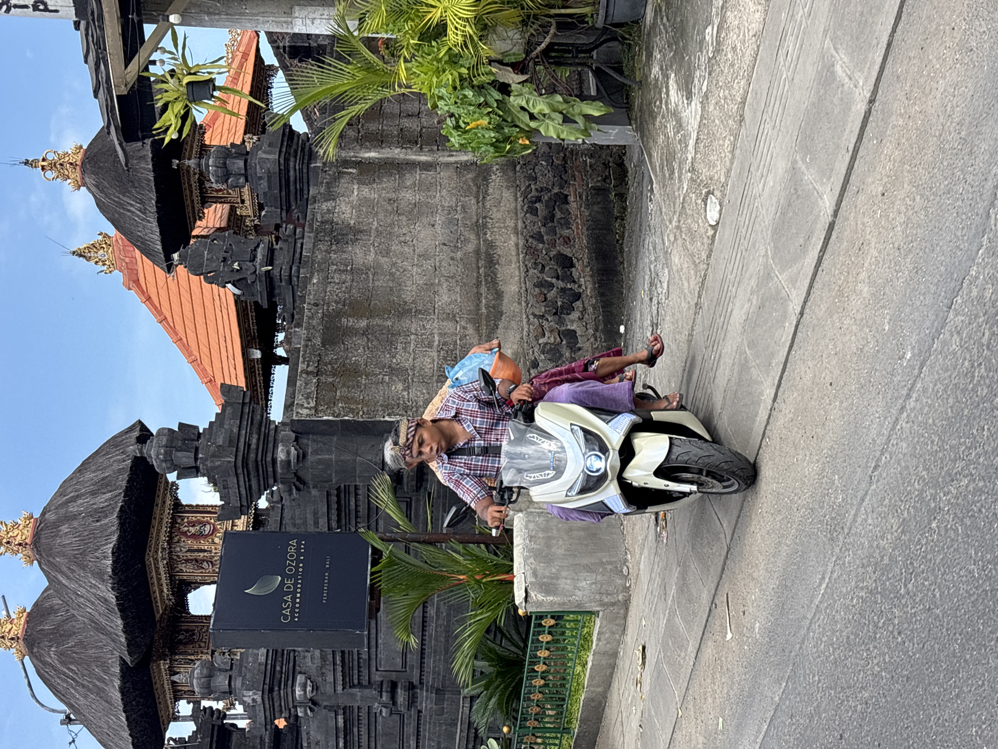 A man is riding a white and black scooter on a paved road in Mengwi, Indonesia. The man is wearing a checkered shirt, light purple pants, and a head covering. A passenger sits behind him, wearing flip-flops. A sign for "Casa de Ozora Accommodation & Spa" is visible next to a traditional Balinese building with a thatched roof. Palm trees and other greenery surround the building. The sky is partly cloudy.