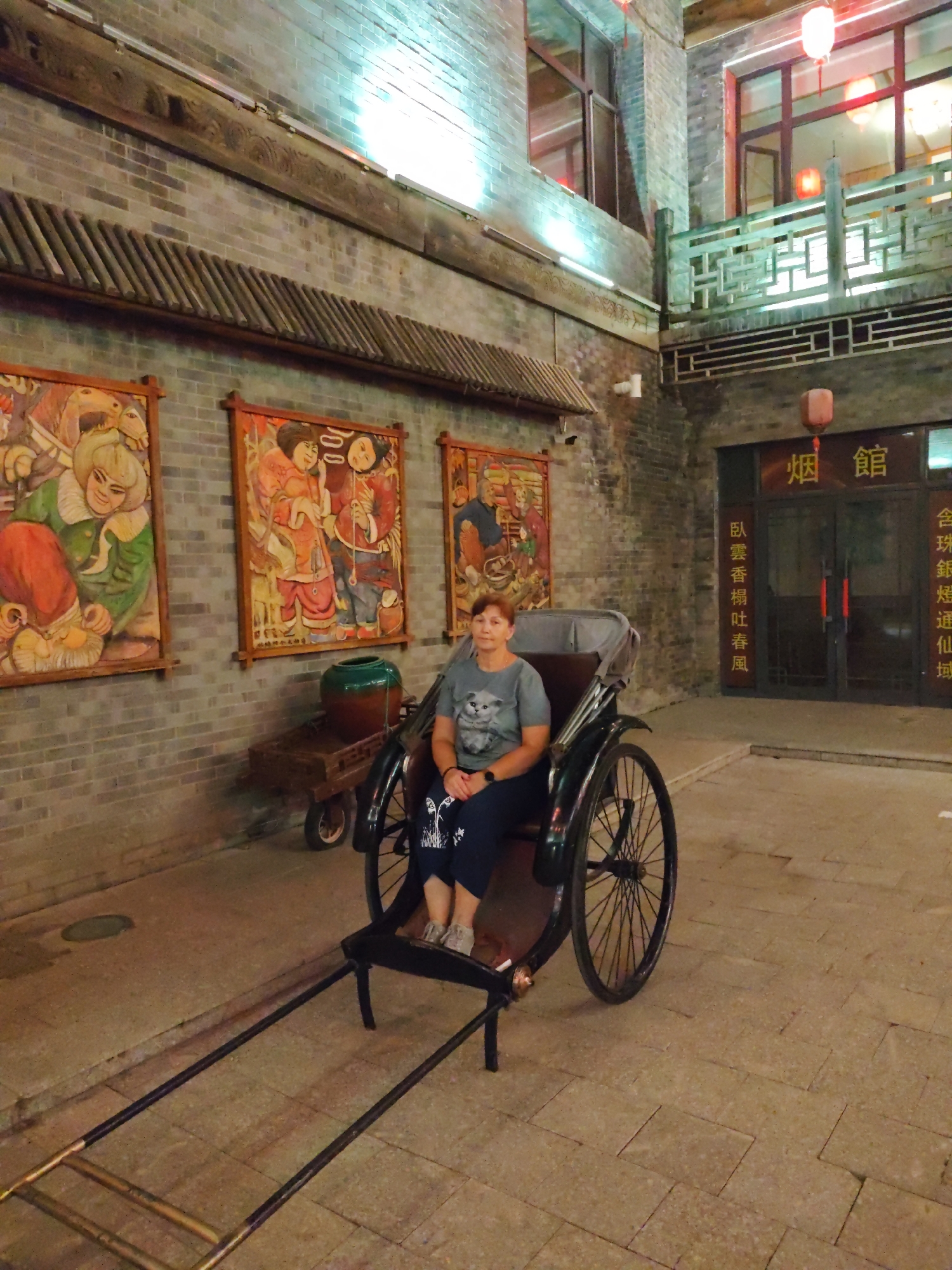 A woman is sitting in a rickshaw in front of a building in Lindian, China. The building has a brick facade and traditional Chinese architecture, including red lanterns and decorative carvings. Three large framed paintings are hung on the wall next to the building. The rickshaw is black with a large spoked wheel at the back and a wooden frame. The woman is wearing a grey t-shirt with a cat print and dark pants. The ground is paved with stone.