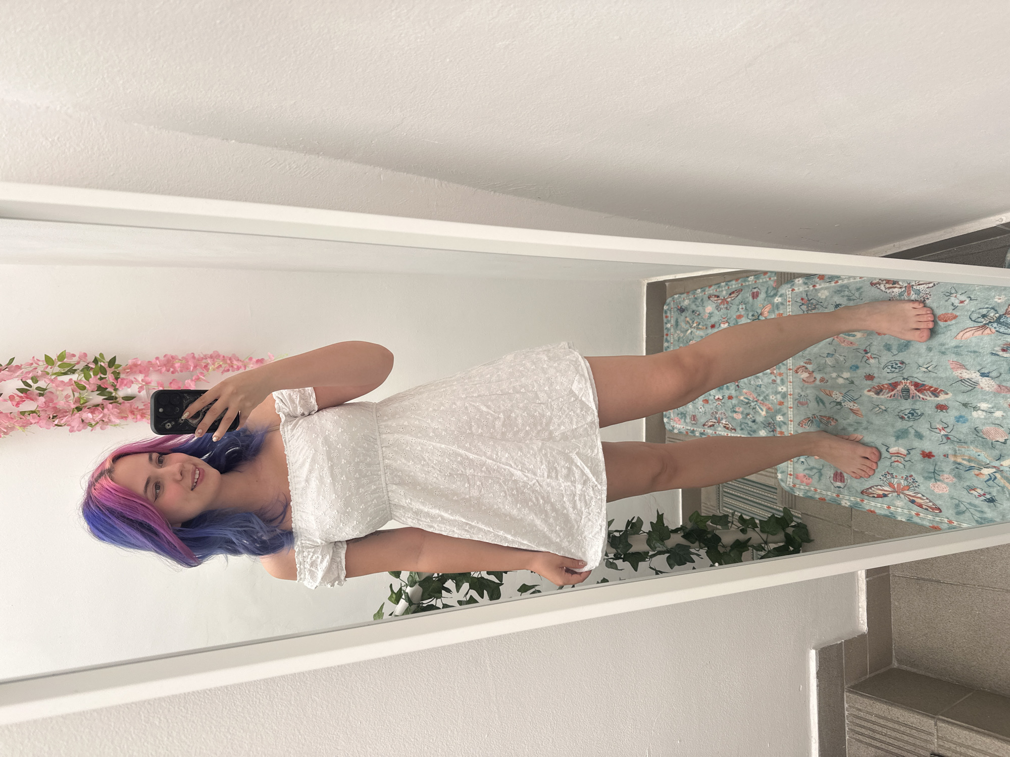A young woman is captured in a mirror selfie, standing indoors and smiling towards her phone. She has distinctive multi-colored hair, with shades of blue and purple, and hints of pink. She is wearing a short, white, off-the-shoulder dress featuring an embroidered or eyelet pattern and a smocked bodice. Her left hand holds a smartphone with a dark, patterned case, positioned to take the picture.
The background behind her includes a plain white wall adorned with a pink floral garland. On the floor, a light blue or teal rug with intricate patterns of butterflies and various insects is visible. Green artificial ivy or vines are also present near the bottom edge of the mirror. The floor beneath the rug appears to be tiled in a neutral color, contributing to the bright, indoor residential setting. The lighting suggests it is daytime. No text is visible in the image.