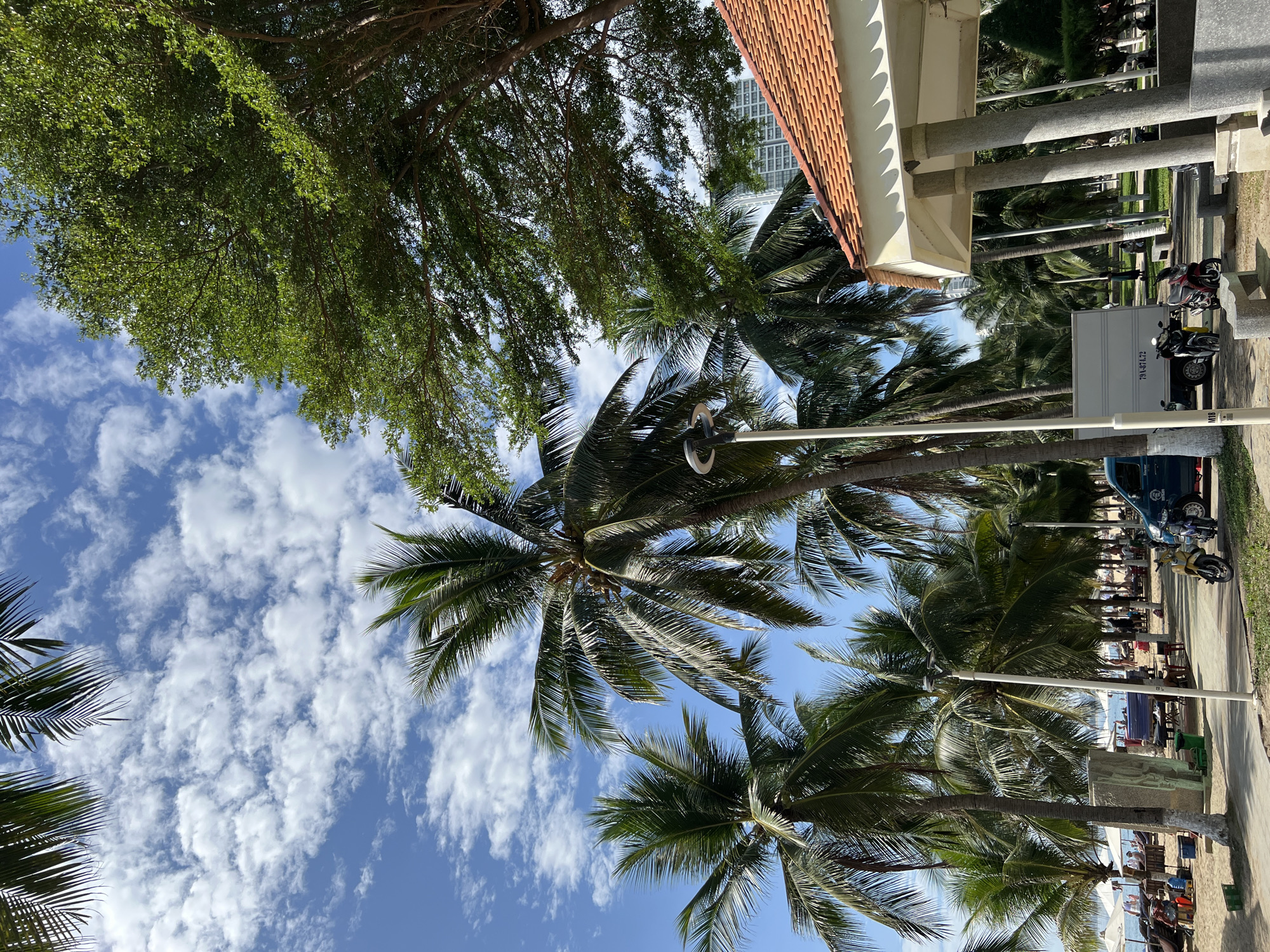 The image depicts a sunny day at a beach in Nha Trang, Vietnam, characterized by palm trees, a clear blue sky with scattered clouds, and a sandy shore. In the foreground, a pathway lined with palm trees leads towards the beach. A small, open-air pavilion with an orange tiled roof stands to the right, supported by concrete pillars. Several motorcycles are parked along the pathway. A white box truck with Vietnamese license plates is also visible. In the background, beachgoers relax on lounge chairs under umbrellas, and the ocean stretches towards the horizon. A tall building can be seen in the distance. The scene conveys a relaxed, tropical atmosphere typical of a beach resort.