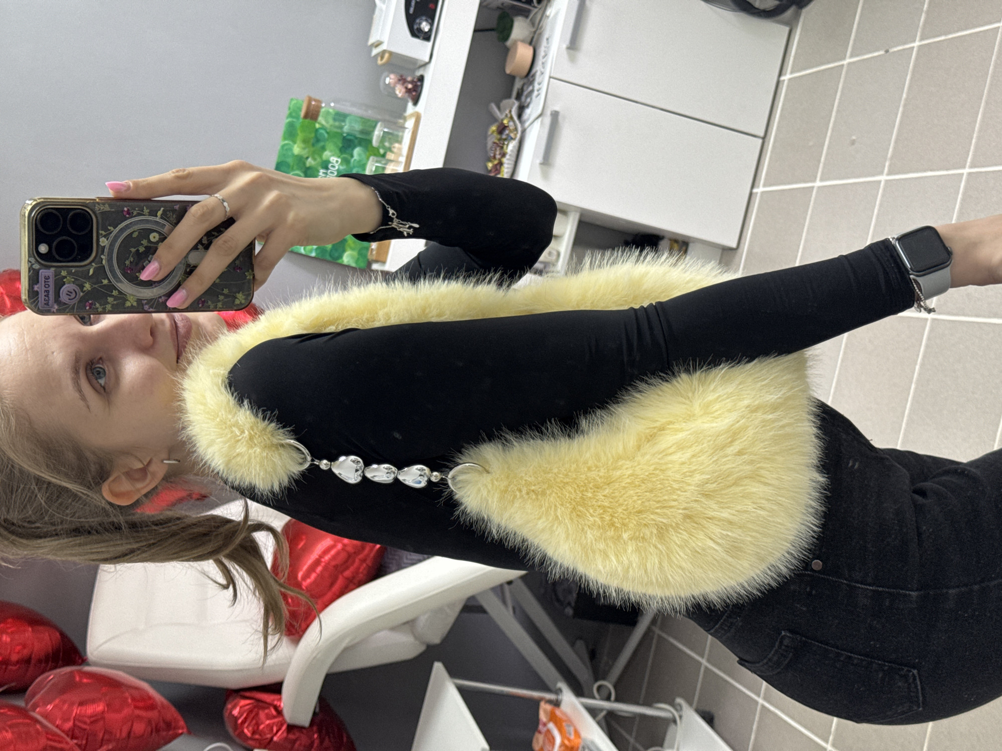 A woman is taking a selfie in what appears to be an indoor setting. She is wearing a black long-sleeved shirt with a yellow fur collar accented by decorative beads. She is holding a phone with a floral-patterned case. She also wears an Apple Watch on her left wrist. The setting includes office-style furnishings such as a white cabinet and a chair, as well as red heart-shaped balloons. The scene is likely in Izhevsk, Russia.