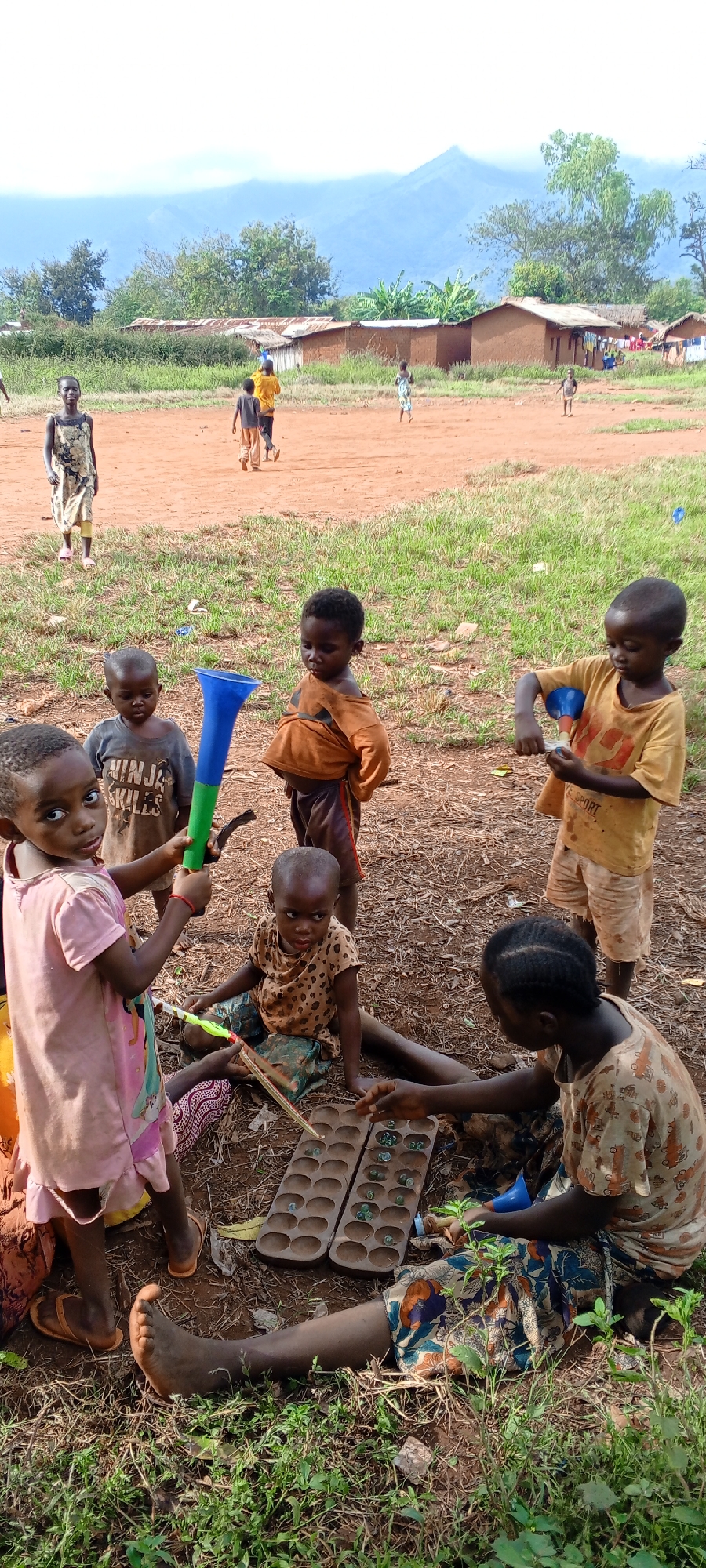The image depicts an outdoor scene in Ngalula, Democratic Republic of the Congo, bustling with children engaged in various activities. In the foreground, a group of young children is gathered on reddish-brown earth interspersed with patches of green grass. An older child or young adult sits on the ground, intently playing a mancala board game with another child. The wooden mancala board features depressions holding small, translucent green game pieces.
Surrounding the game, other children interact. A young girl in a light pink dress stands, holding a blue and green plastic funnel, looking directly at the camera. Next to her, a boy wears a grey t-shirt with the text "NINJA SKULES" visible. To the right, a boy in a yellow t-shirt, bearing the word "SPORT," holds a blue and yellow plastic funnel, seemingly examining something in his hand.
In the mid-ground, several other children walk across an open, unpaved area. Behind them, simple houses constructed from reddish mud bricks and topped with corrugated iron roofs are visible. Lush green trees and vegetation form a natural backdrop around these dwellings. Distant, hazy mountains rise under a bright sky in the far background.
The scene portrays a lively and communal atmosphere of children playing and socializing freely outdoors. The time of day appears to be daytime, possibly late morning or early afternoon, with clear but potentially hazy weather. The ground is dry, characteristic of an unpaved village setting. The children's clothing is simple, consistent with a rural environment.