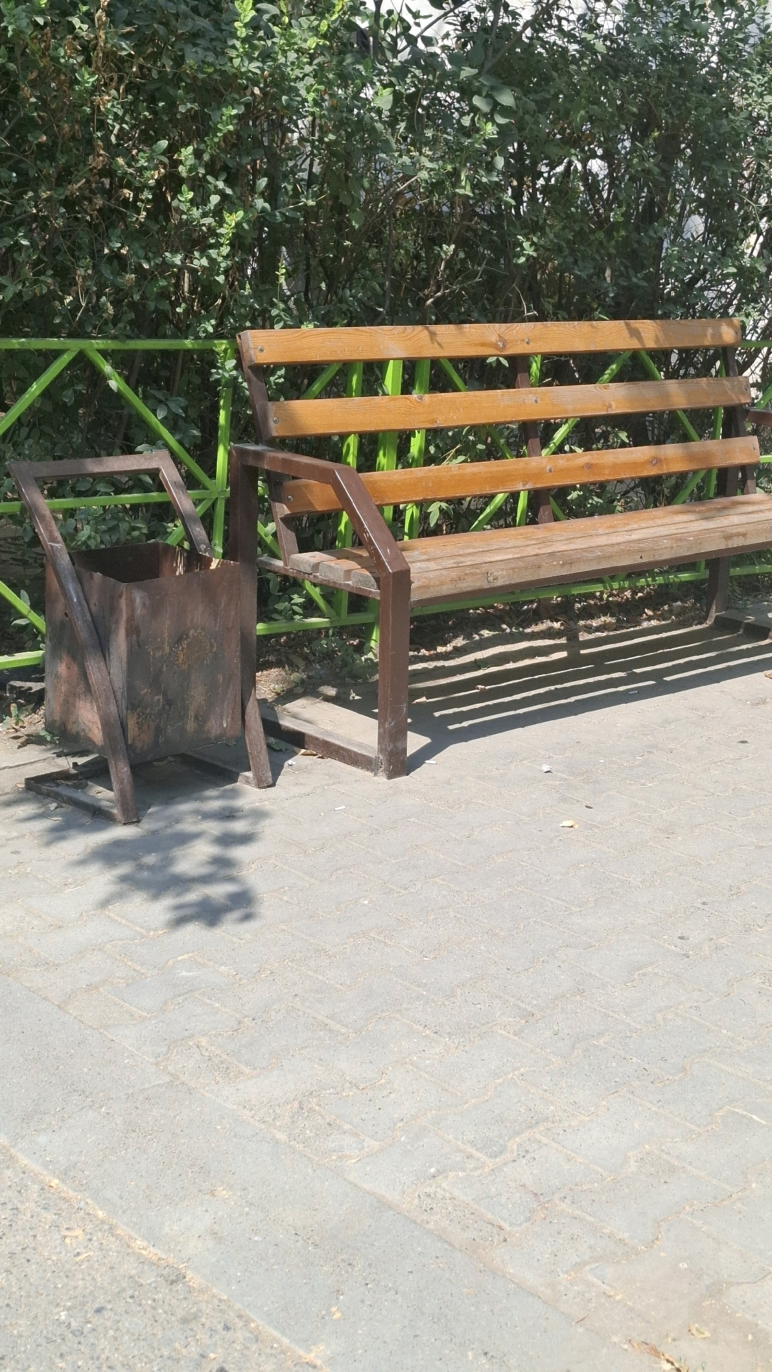 An outdoor scene depicts a park bench and a rusty metal trash can on a paved walkway. The bench is made of wooden slats supported by a dark metal frame, and it is positioned in front of a dense green hedge and a green metal fence. The trash can, made of weathered metal, is to the left of the bench, with its opening facing forward. The ground is covered with interlocking pavers, and shadows from overhead foliage are cast on the walkway. The image appears to be taken in Shymkent, Kazakhstan, likely a public space such as a park or a street. No people are visible in the frame.