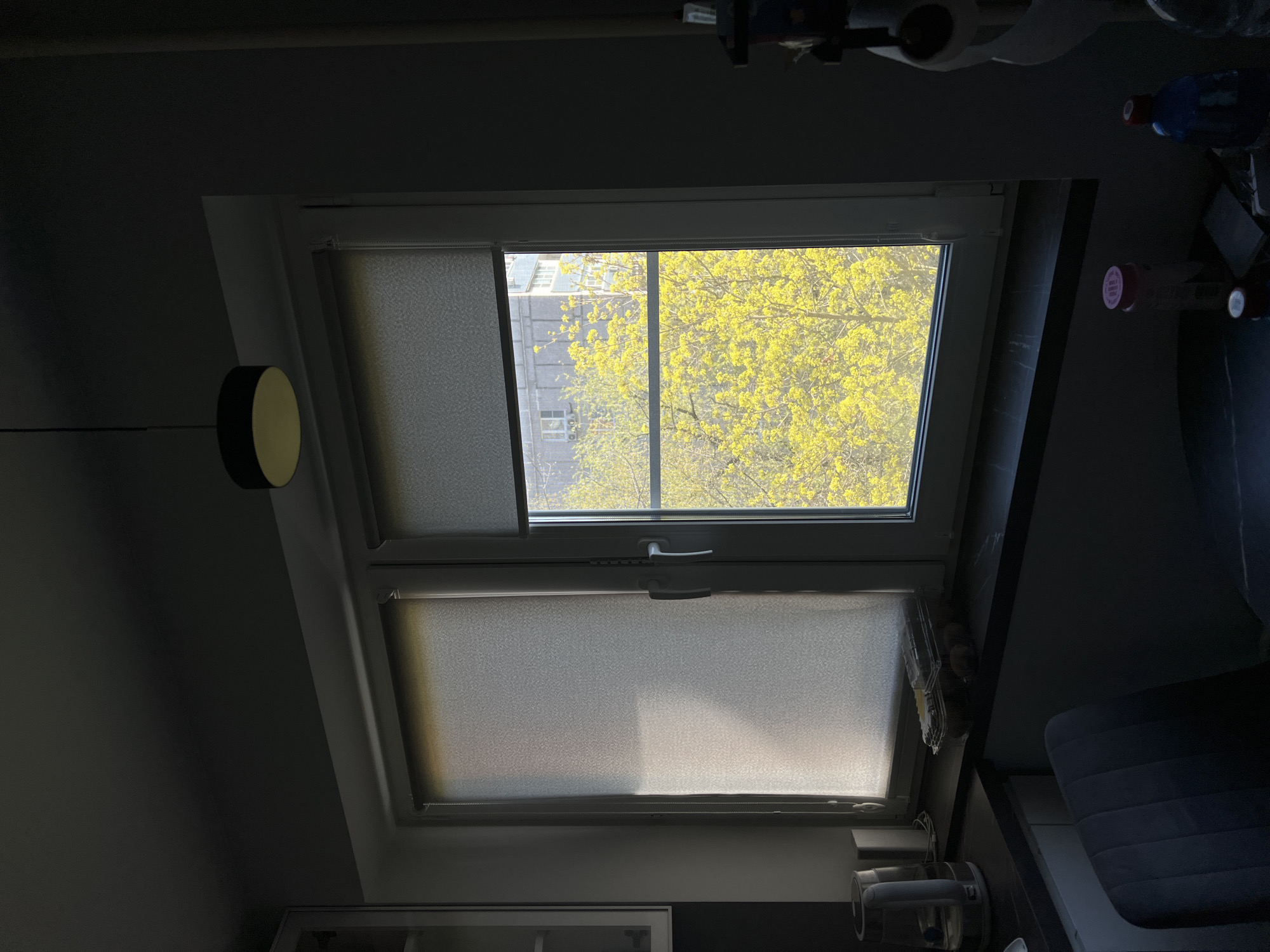 The image shows an interior view of a room with a window. The window is divided into two sections. The top section has a partially lowered roller blind on the left side, while the right side is open, revealing the view of trees with yellow leaves outside, possibly indicating springtime. The bottom section of the window also has a roller blind, which is partially lowered. The room appears to be dimly lit, with dark walls and furniture. A round, flat lamp is visible to the left of the window. In the background, above the window, there are some unidentified objects and containers. The location is unknown.