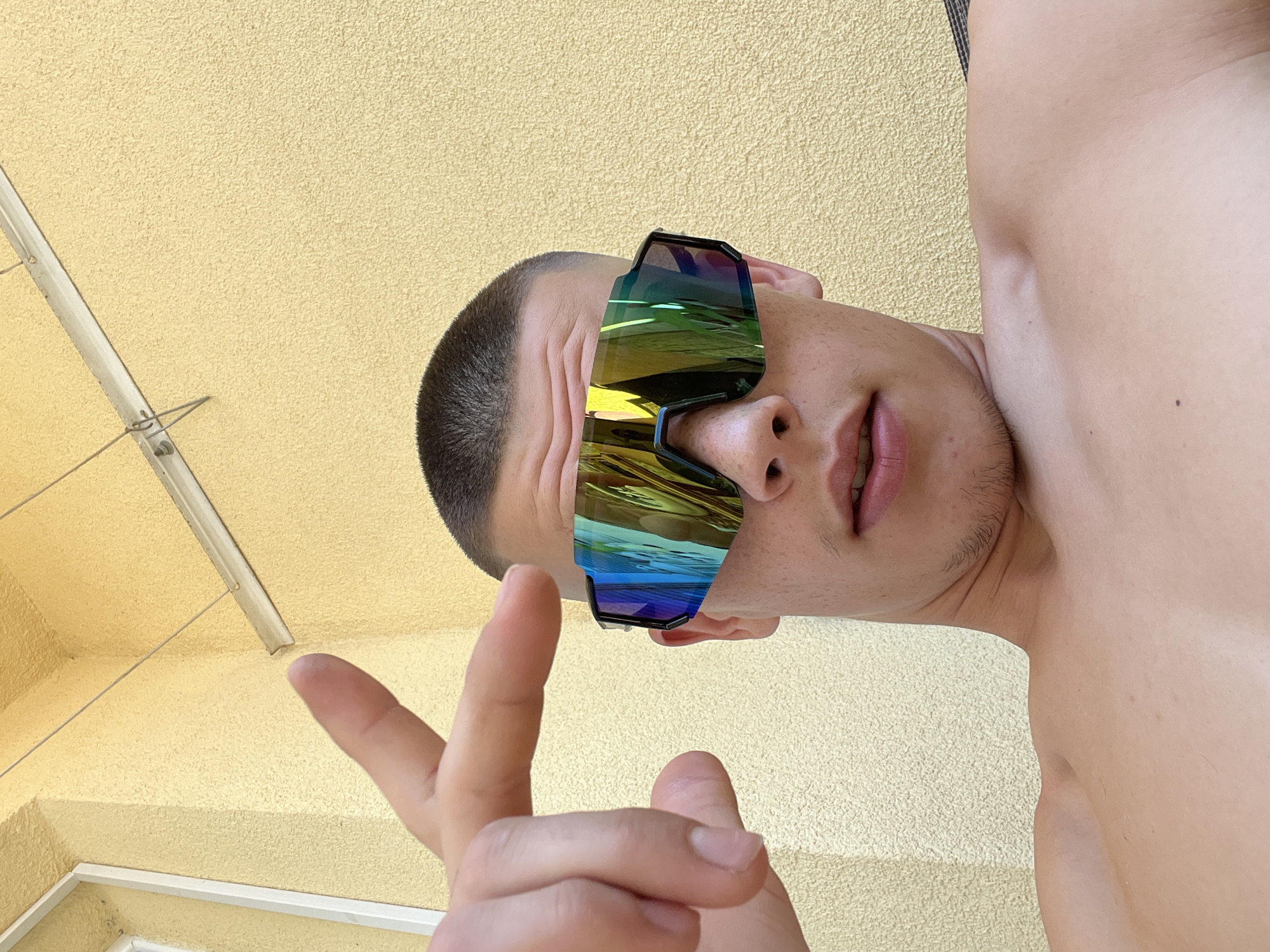 An outdoor close-up, selfie-style shot shows a young Caucasian male with a buzz cut and a shirtless torso. He is wearing large, reflective, multi-colored wraparound sunglasses. His left hand is raised, with his index finger and thumb extended in a gesture. The background is a textured, pale yellow wall with a light-colored metal fixture and thin wires running across it. The lighting suggests a bright, sunny day. The scene does not provide any specific information about the city of Sopot, Bulgaria.