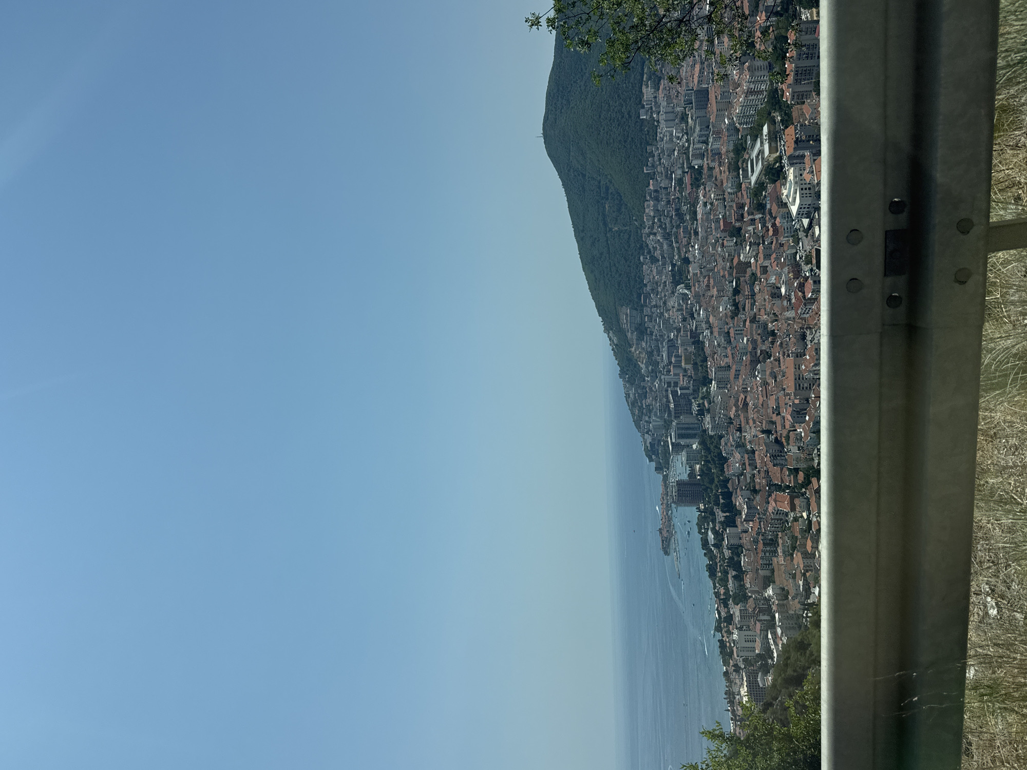 An elevated, medium-shot captures a sprawling coastal city nestled between a verdant, tree-covered mountain and the calm, blue sea under a clear sky. In the foreground, a weathered, gray metal guardrail runs horizontally across the frame, with dry grass visible below it. The city itself is a dense collection of buildings, many with terracotta-tiled roofs, that extend up the gentle slope of the mountain. Modern high-rise buildings are interspersed with older, lower-rise structures. A harbor with several boats is visible along the coastline. The mountain behind the city is heavily forested, with a single structure, possibly a tower, visible at its peak. The overall scene suggests a picturesque, Mediterranean landscape on a bright, sunny day.