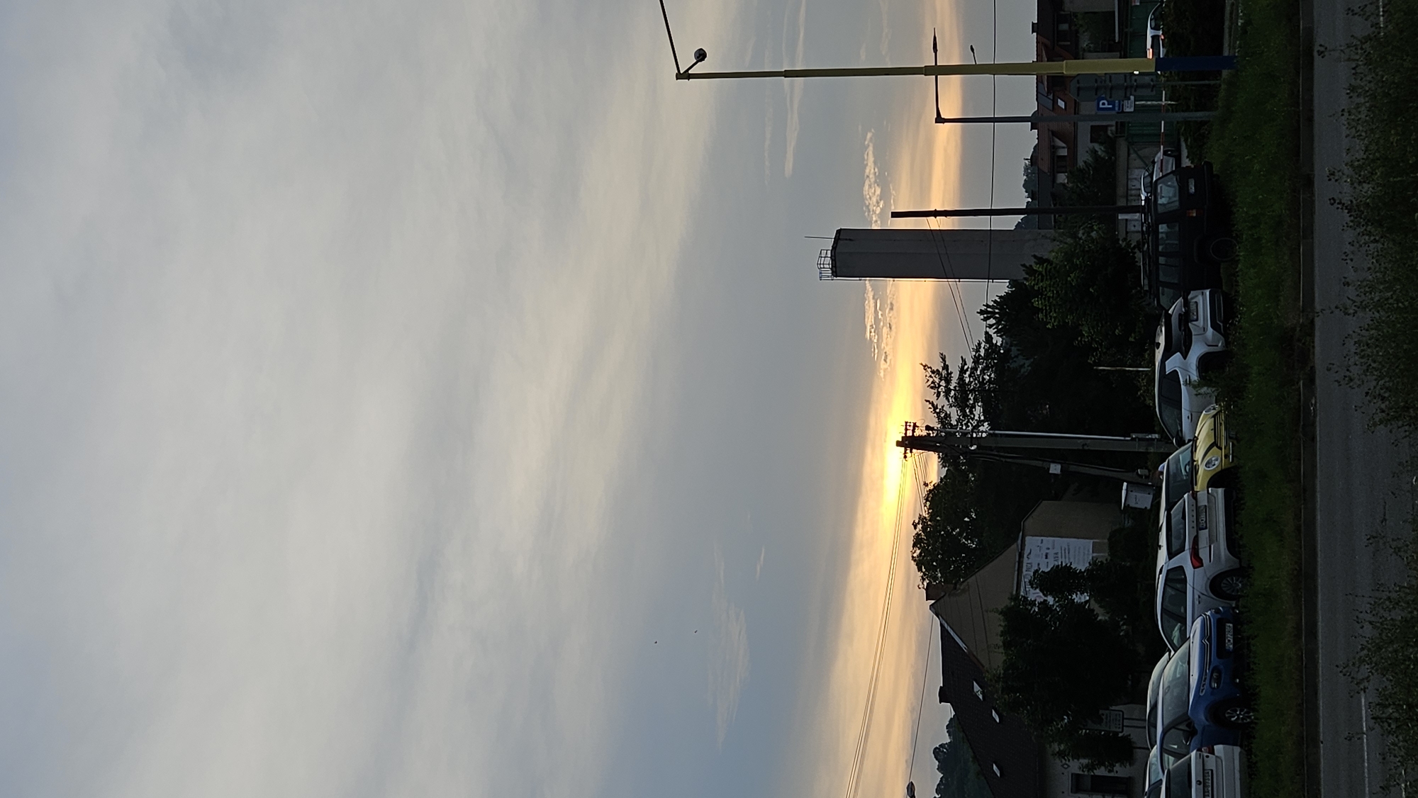 The image depicts a rural street scene in Prešov, Slovakia, during what appears to be sunset. The sky is filled with soft, wispy clouds that are illuminated by the warm hues of the setting sun, casting a golden glow. The overall mood is peaceful and serene.
On the right side of the image, a row of parked cars is visible, mostly compact SUVs and sedans. These vehicles are parked along a grassy verge next to a paved road. Behind the cars, a line of trees adds a touch of greenery to the scene. In the background, a tall, rectangular structure, possibly a water tower, stands against the illuminated sky. Further back, houses with pitched roofs are nestled amongst the trees, suggesting a residential area.
Several utility poles and wires are also present, indicating the presence of infrastructure in the area. One pole, in particular, has a lamp attached to it, suggesting it is a street light. The light from the setting sun creates a dramatic contrast between the illuminated sky and the silhouetted elements of the landscape.
The image is captured vertically, which emphasizes the height of the structures and the expanse of the sky. There are no people visible in the frame. The weather appears to be clear, with the exception of the scattered clouds. The time of day is clearly indicated by the dramatic colors of the sunset. There is no visible text in the image.