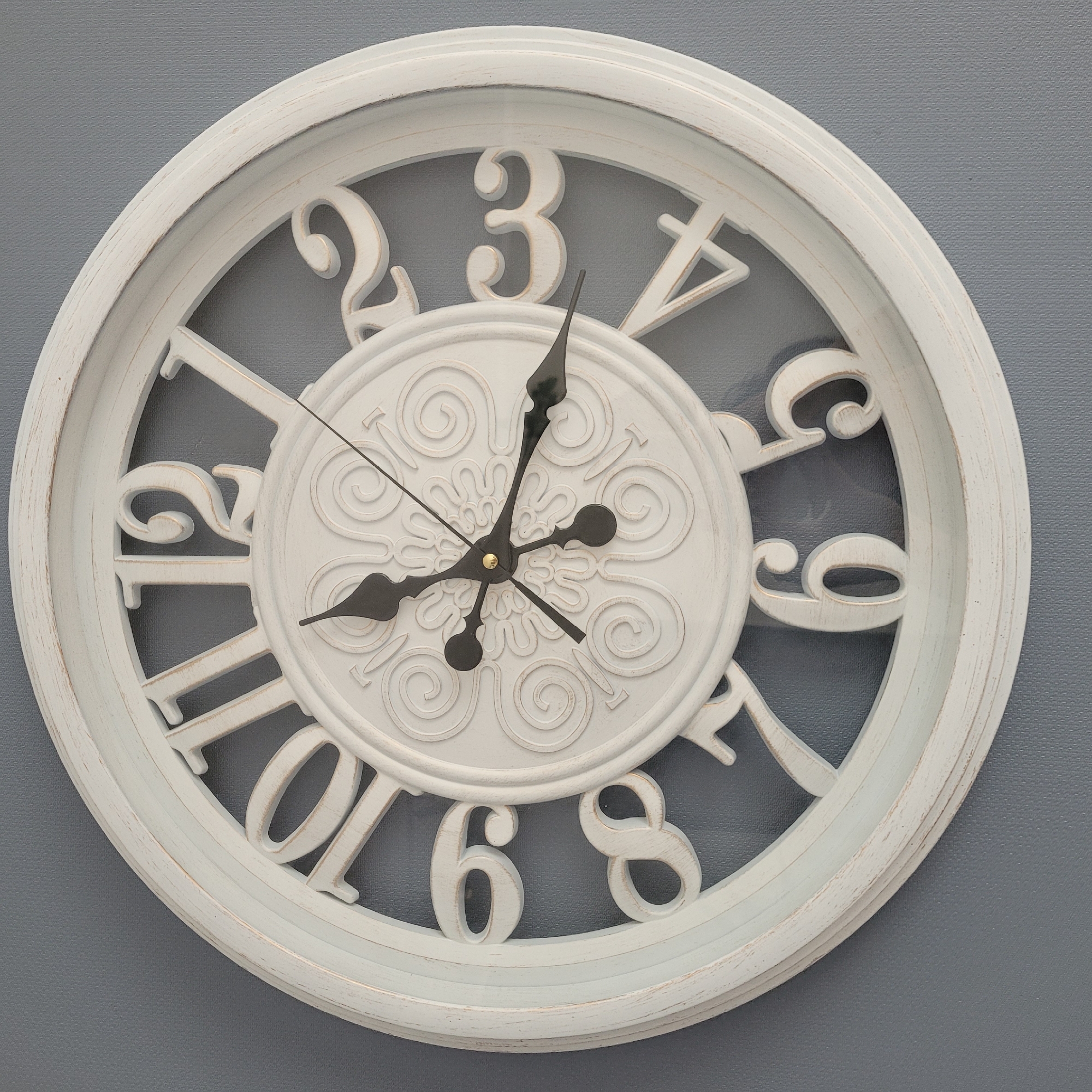A close-up, eye-level, studio shot showcases a round, white, decorative wall clock with Roman numerals against a textured gray wall. The clock has a distressed finish, with hints of the wood beneath showing through the white paint, particularly on the edges and the raised numerals. The clock face features an ornate, embossed circular pattern at its center. Three black hands are visible: a short hour hand pointing slightly past the 10, a long minute hand pointing at the 2, and a thin second hand extending towards the 4. The clock is centrally positioned in the frame. This image was taken in Jūrmala, Latvia.