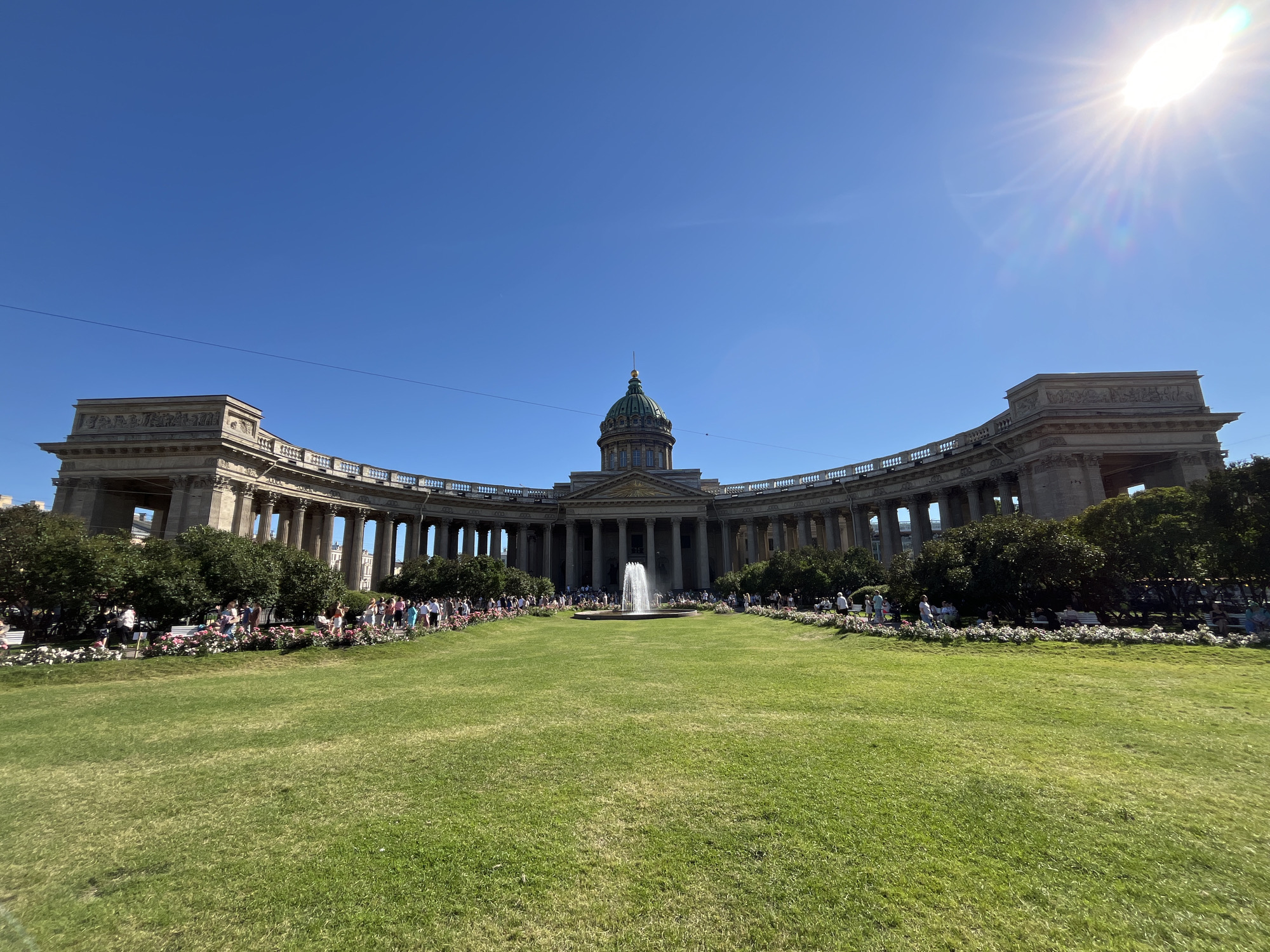 This image depicts a wide-angle view of Kazan Cathedral in Saint Petersburg, Russia, on a clear, sunny day. The cathedral, an iconic architectural landmark, features a grand semicircular colonnade with numerous Corinthian columns leading to a central dome. In front of the cathedral, a lush green lawn stretches outwards, bisected by a pathway lined with flowering bushes and trees. A fountain sprays water in the center of the lawn, adding a dynamic element to the scene. A crowd of people is visible strolling along the pathway and gathering in front of the cathedral, suggesting it is a popular tourist destination or public space. The sky is a vibrant blue, with the sun positioned in the upper right corner, casting a bright glare and lens flare across the image. The overall scene conveys a sense of grandeur and vibrant activity in an urban setting.