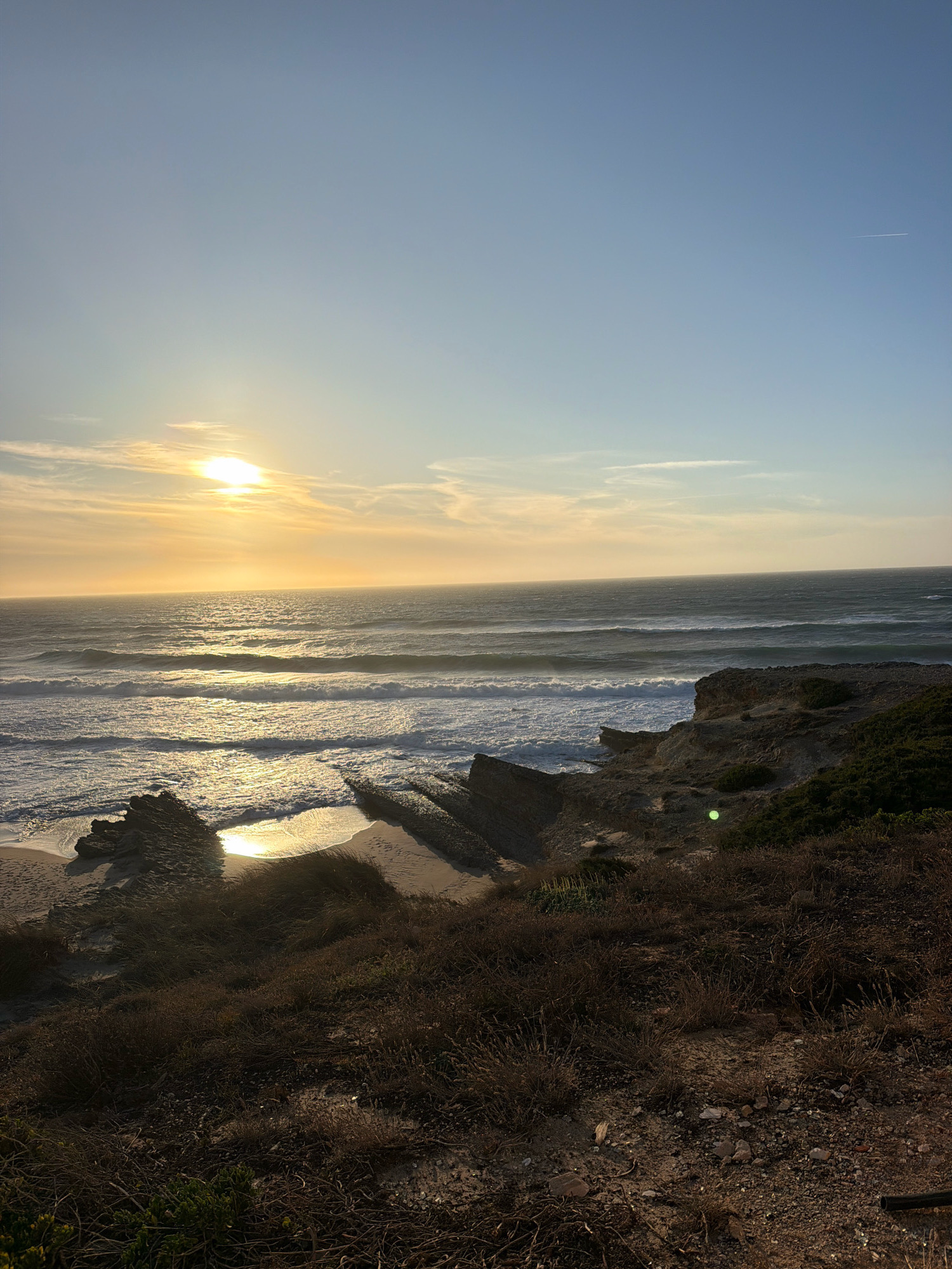 This is a landscape photograph of a coastal scene at sunset in Cascais, Portugal. The image is dominated by the ocean, with waves breaking on a sandy beach and rocky outcrops. The sun is setting on the horizon, casting a warm glow across the water and sky. The foreground is covered in dry vegetation and sand, suggesting a natural, undeveloped area. The sky is a clear blue with a few wispy clouds. The overall impression is one of peace and natural beauty.