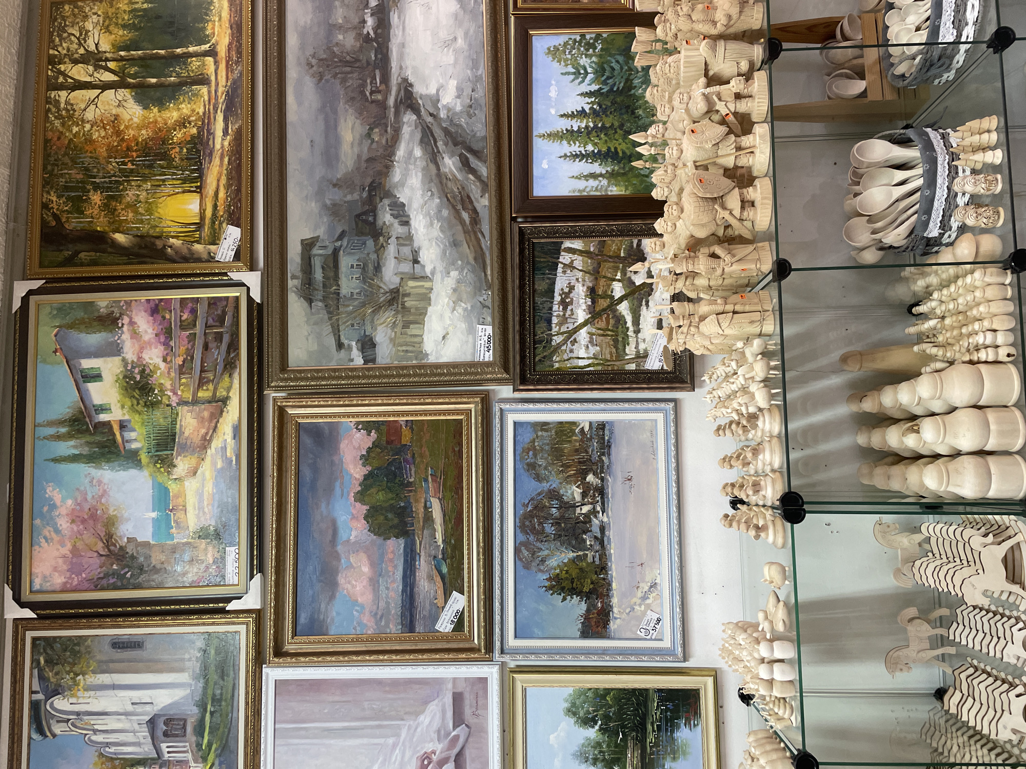 The image shows a display of traditional Russian crafts and artwork in what appears to be a shop or market stall in Sergiyev Posad, Russia.
The subjects are primarily wooden handicrafts and paintings. On the left side of the image, several framed paintings are displayed, depicting landscapes and architectural scenes. These paintings showcase varied artistic styles, from realistic to more impressionistic.
The main focus of the display, particularly on the right side, is an assortment of intricately carved wooden items. These include chess pieces, possibly representing historical figures or knights, as well as wooden spoons and other decorative carvings. The wooden items are arranged on glass shelves, suggesting a retail environment. Some of the chess pieces are quite elaborate, with detailed armor and weaponry. The wooden spoons are bundled together in baskets or containers, some with decorative trim.
The overall setting is a bright indoor space, likely a shop. The lighting highlights the textures and details of the wooden crafts and the colors of the paintings. There is no discernible activity or event taking place; the scene appears to be a static display for viewing and purchase.
Notable details include price tags attached to some of the paintings, with visible text such as "45.000" and "18 000". This indicates the items are for sale. The style of the paintings and the traditional wooden crafts strongly suggest a Russian cultural context, consistent with the provided location. The atmosphere seems calm and inviting, typical of a place showcasing artisanal goods.