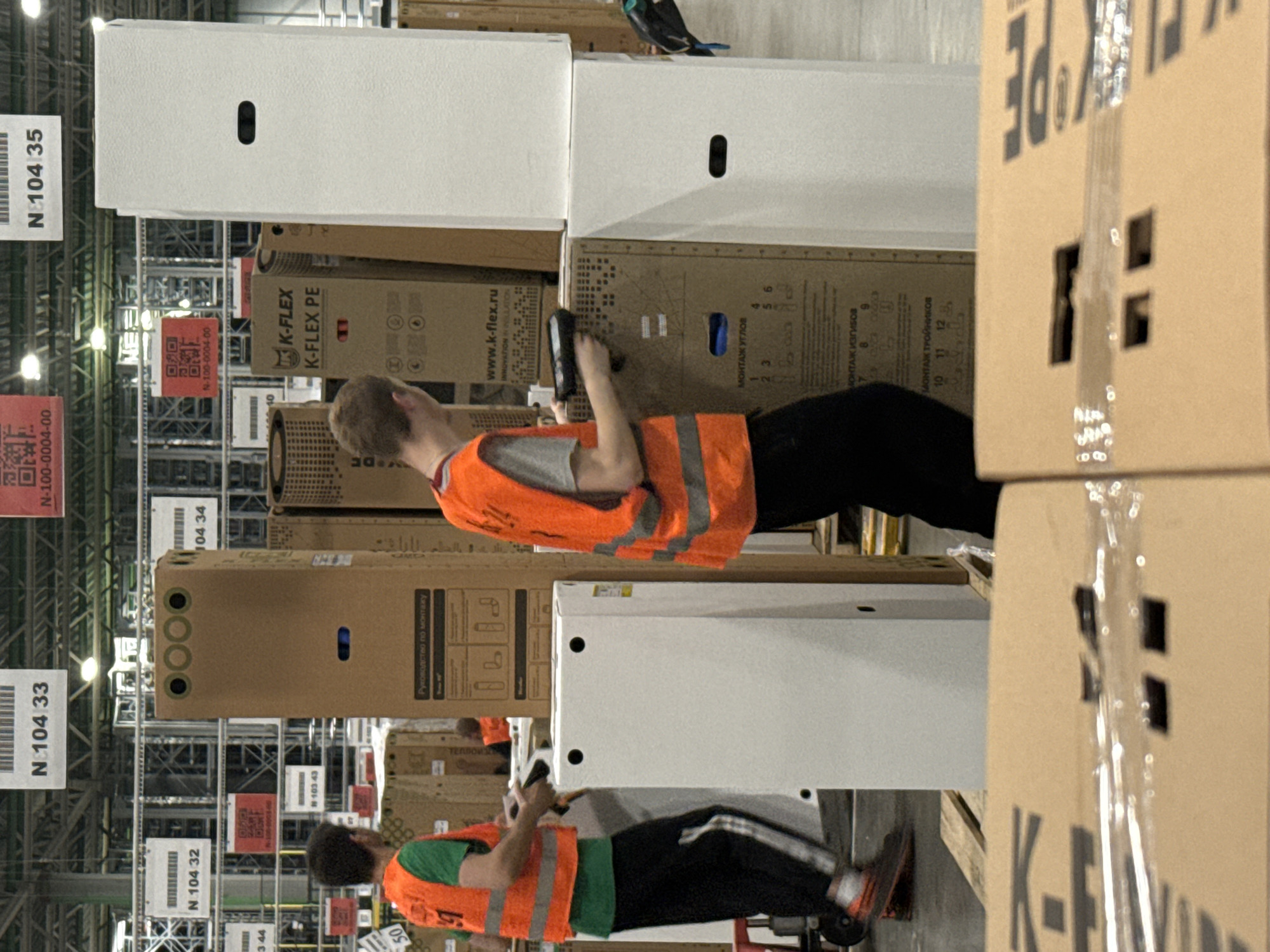 The image depicts an indoor warehouse environment, likely a logistics or distribution center, in Lupanovo, Russia. Two individuals wearing orange high-visibility vests are present. One individual, on the left, is holding a handheld scanner and appears to be scanning an item. The other individual, on the right, is partially obscured and also wearing an orange vest. Both individuals are in proximity to large cardboard boxes and white rectangular objects, possibly appliances or equipment. Overhead, signage with alphanumeric codes, such as "N 104 33" and "N 104 32," is visible, suggesting a system of organization and inventory management. The lighting appears to be artificial, consistent with an indoor industrial setting. The floor is concrete.