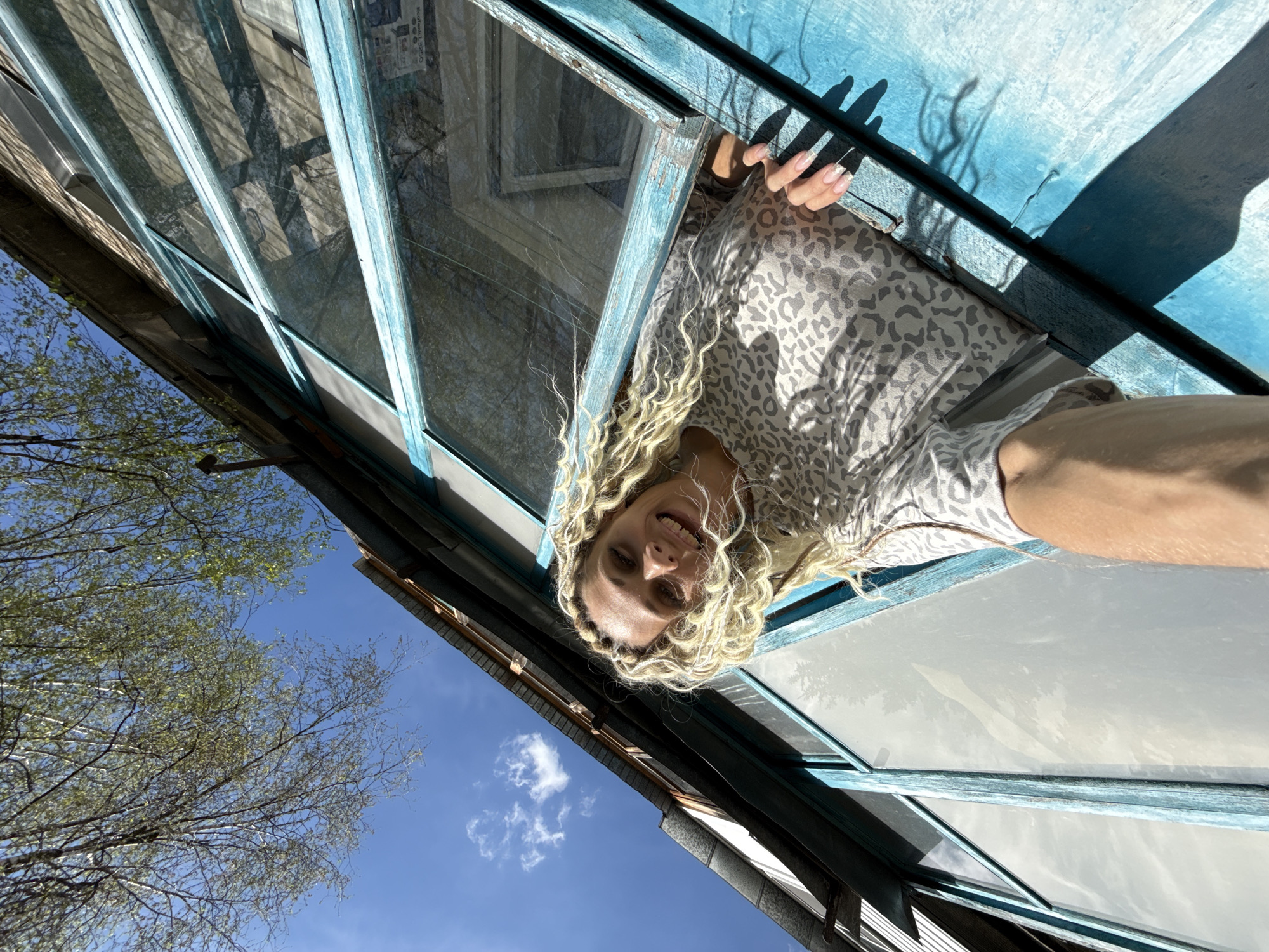 A woman with blonde curly hair is looking into the camera from above, standing outside a structure in Rusanovo, Russia. She is wearing a leopard-print shirt. The structure appears to be a building or greenhouse with windows framed in light blue paint. The camera is positioned at an angle, capturing the blue sky, branches of a tree, and a small cloud in the background. Part of the building's exterior is visible. The woman's hand with painted fingernails is visible on the window frame.