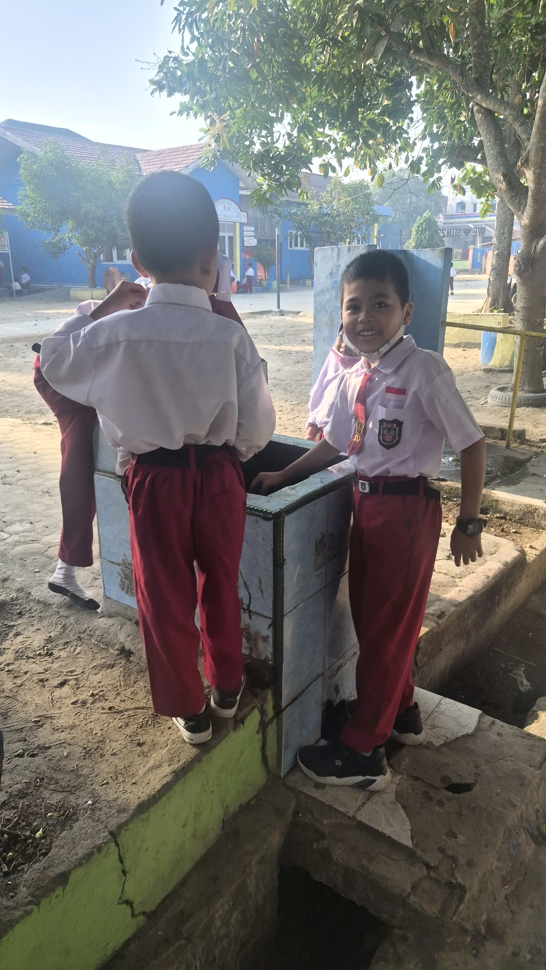 Two young boys, likely students, are standing near a tiled structure in a schoolyard. The boy on the right is wearing a white collared shirt, red pants, a red tie with a school emblem, and a watch on his left wrist. He is smiling and looking at the camera. The boy on the left is wearing a white collared shirt and red pants, facing away from the camera. Behind them, a blue school building with a red roof is visible, surrounded by trees and vegetation. The ground is dirt and concrete. The overall impression is a sunny day at a school in a tropical climate. The location is identified as Putak, Indonesia.