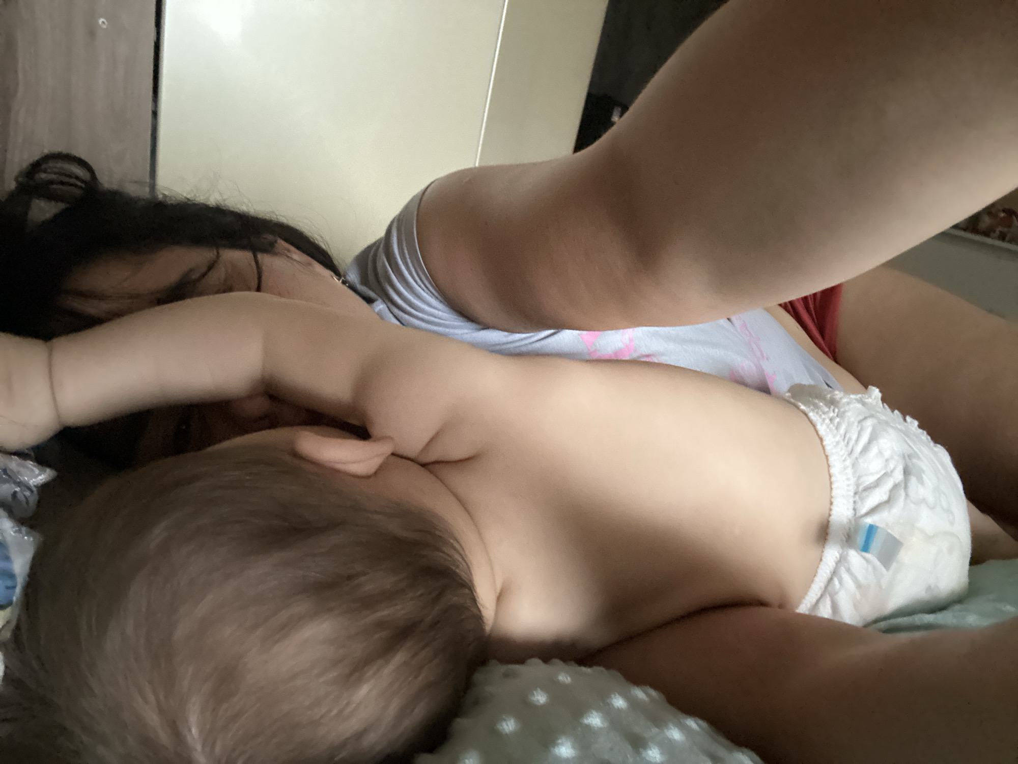 A woman with dark hair is lying down while a baby wearing a diaper lies on top of her. The woman is wearing a gray shirt with a pink design, and possibly red shorts or underwear. The baby is facing away from the camera. The scene appears to be indoors, possibly in a bedroom. The location is Unknown, Unknown.