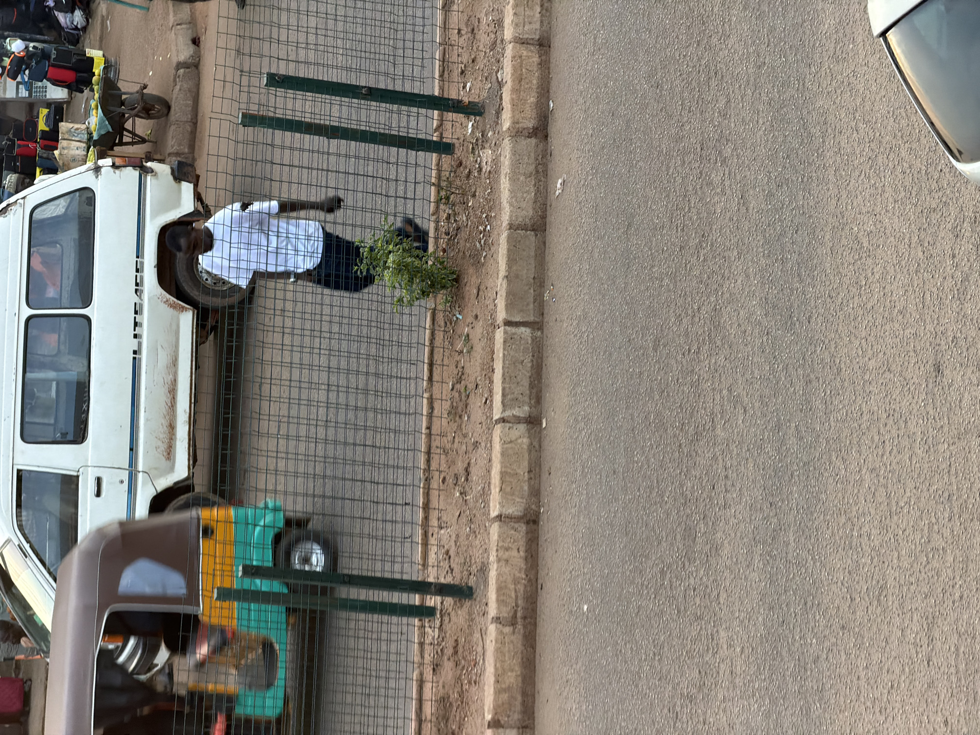 In Bukuru, Nigeria, a man wearing a white t-shirt and dark trousers walks behind a chain-link fence. To his left, a three-wheeled auto-rickshaw, predominantly brown with green and yellow accents, is in motion. Behind the man, a white van with blue trim is parked on the street, and a wheelbarrow laden with produce sits on the sidewalk. The ground is paved with asphalt, and a concrete curb separates it from a dirt area where the fence is situated.