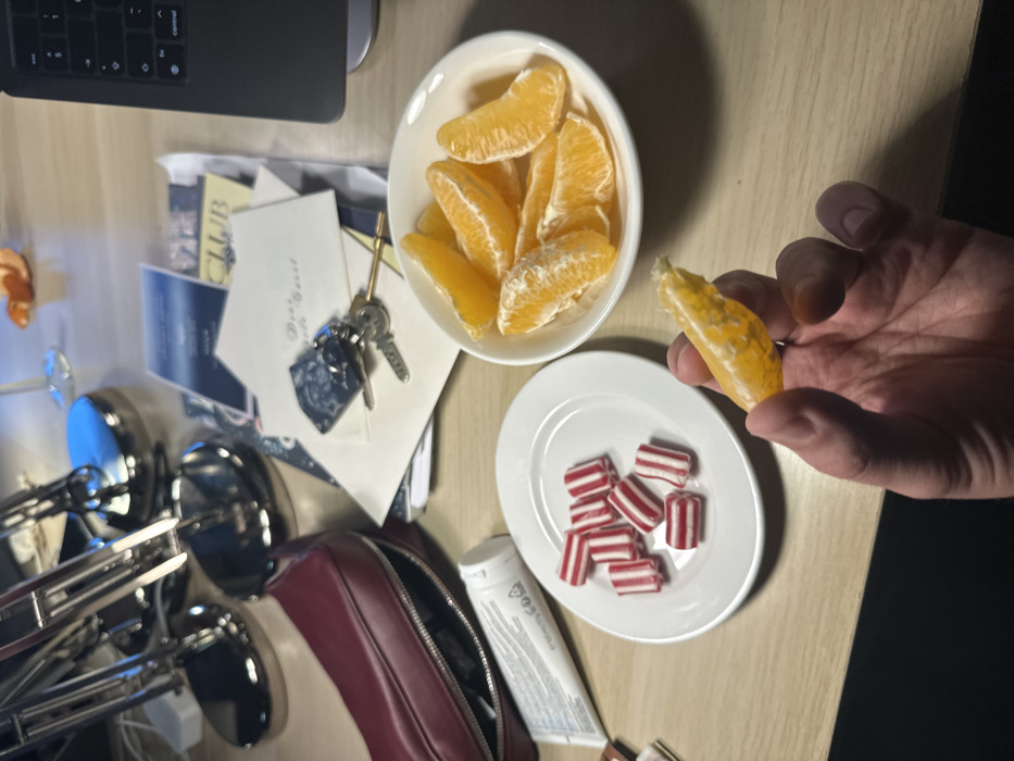 An overhead shot reveals an assortment of items on a table. A bowl of orange segments sits adjacent to a small plate holding red and white striped candies. A hand holds an orange segment. Nearby are keys atop some papers, a makeup bag, and a tube of cream. A desk lamp and a laptop keyboard are partially visible in the periphery.