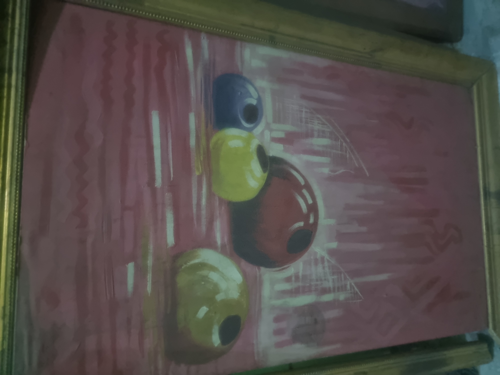 The image depicts a framed painting with a gold frame. The painting has a reddish-pink background with vertical brushstrokes and a zig-zag pattern. The main subject of the painting consists of four colored spherical objects stacked vertically: blue, yellow, red, and yellow again, each with a black circular "hole". White abstract lines and geometric shapes are also visible in the background. The location in Gwagwalada, Nigeria, is not discernible from the image alone. There are no people visible in the image.