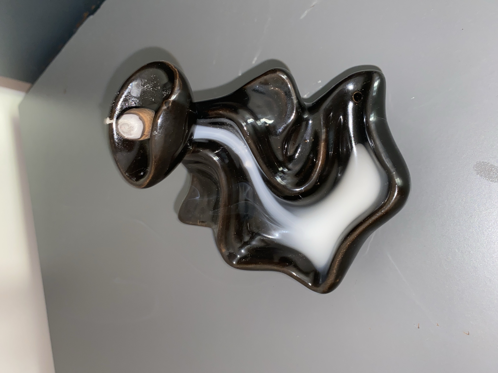 The image shows a brown and white ceramic ashtray on a grey surface. A partially smoked cigarette or joint rests in a holder on one side of the ashtray. The ashtray has a swirling design. The background is out of focus, revealing a white wall and a blue object.
There is no information to suggest that the image has any connection to the city of Krasnoyarsk, Russia.