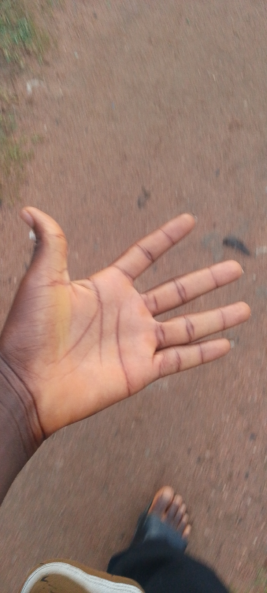 The image shows a person's hand with its palm facing upward and fingers slightly spread. The skin tone is dark, and prominent lines are visible on the palm. The background is a blurry, reddish-brown surface, suggesting an outdoor environment. In the lower right corner, a foot wearing a black sandal is visible, indicating the perspective is from a standing or walking individual. The overall scene appears to be an everyday outdoor setting, possibly a street or path, in Jauro Yinu, Nigeria.
