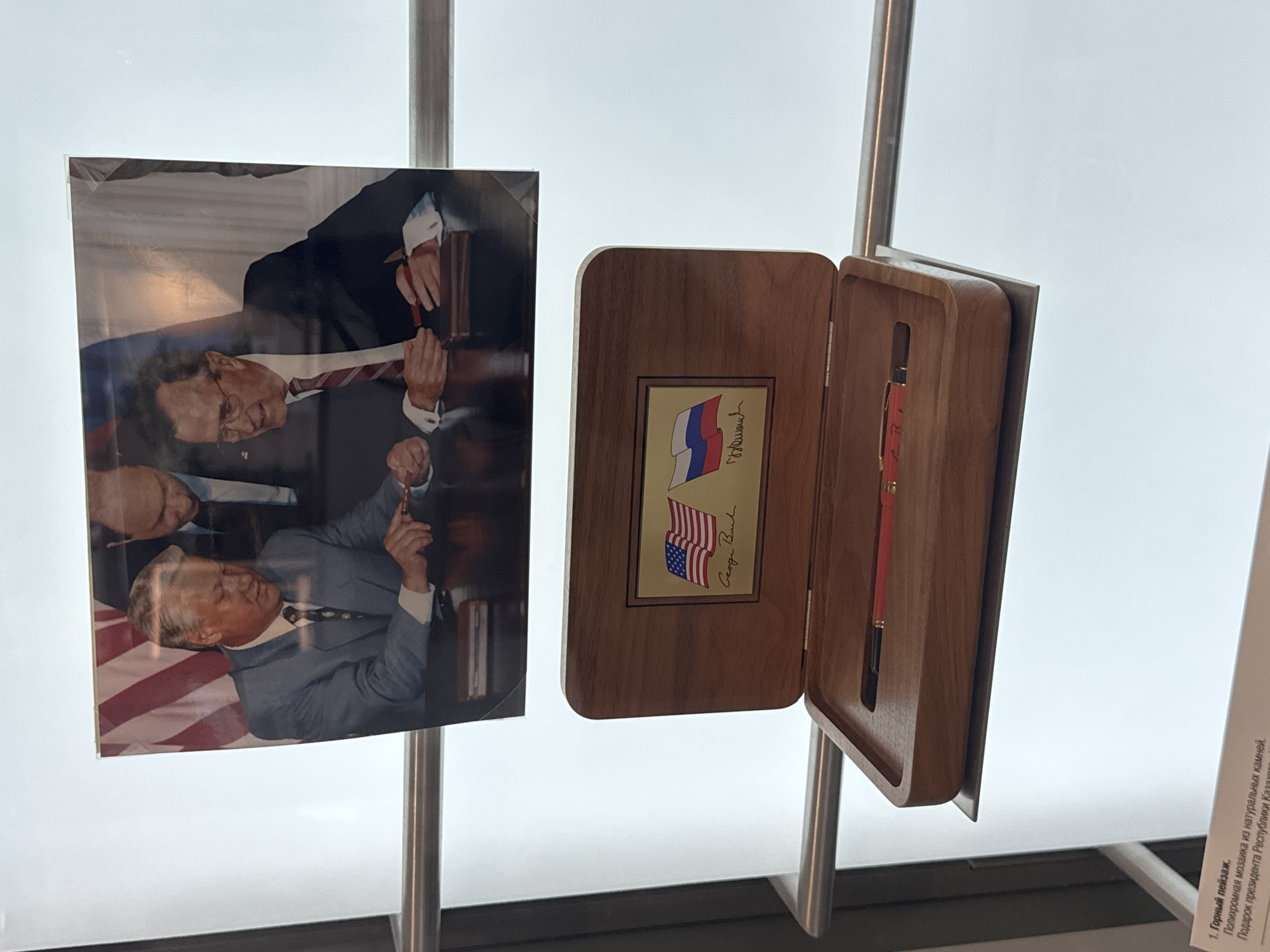 The image shows a display in Yekaterinburg, Russia, featuring a framed photograph of two men signing a document, and a wooden box containing a pen. The photograph, positioned above the box, depicts two figures in suits at a table, with a partial view of the American flag in the background. The men are engaged in signing. Below the photograph, a wooden box is open, revealing a red pen resting in a carved compartment. The lid of the box has a brass plate with the American and Russian flags and two signatures, one appearing to read "George Bush" and the other "Yeltsin". A label at the bottom of the image in Russian text mentions "Polykhromnaya mosaic from natural stones" and "Gift from the President of the Republic of Kazakhstan". The display is likely located in a museum or exhibition space in Yekaterinburg, given the presence of the Russian text and cultural artifacts.