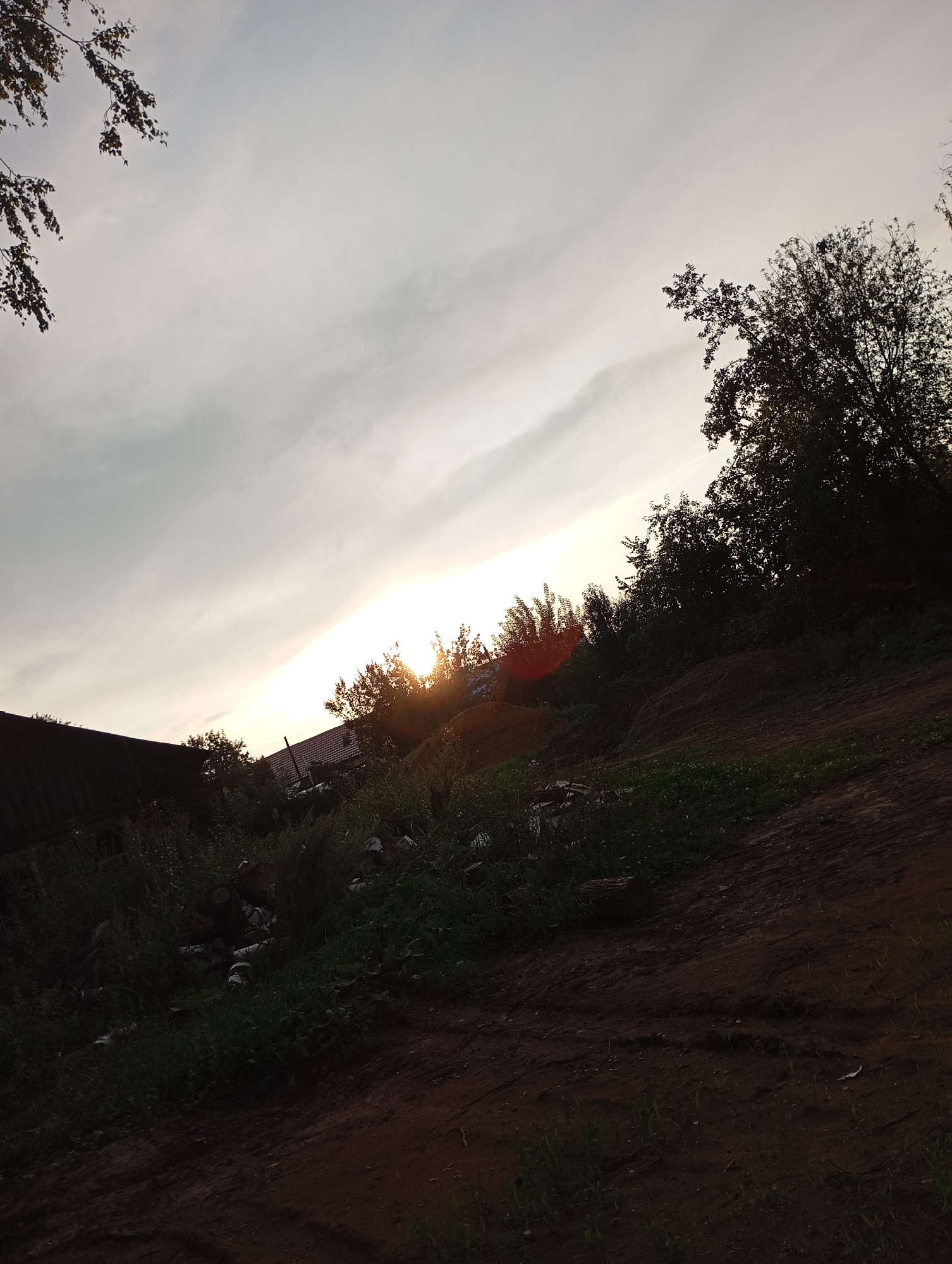 The image depicts an outdoor scene with a diagonal composition, likely taken from a low angle. The sky occupies the upper half, featuring a cloudy expanse with the sun peeking through a gap in the clouds, creating a bright, hazy area. In the foreground and midground, there is a sloped embankment covered in dark earth and sparse green vegetation. A dark, shadowy area on the left side of the frame reveals the edge of a dark-colored building, possibly a shed or a garage, with a dark roof. Further to the right, obscured by trees and vegetation, are rooftops of what appear to be houses. A cluster of dark green trees and bushes dominate the right side of the image, with their branches silhouetted against the bright sky. Some scattered debris, including logs and possibly white plastic items, are visible amidst the overgrown grass in the lower left quadrant. The overall impression is of a rural or semi-rural setting, possibly on the outskirts of the city of Фоки, Russia, during daylight hours, with the sun low in the sky, suggesting either sunrise or sunset. There are no people visible in the image, and no specific actions or interactions are depicted.