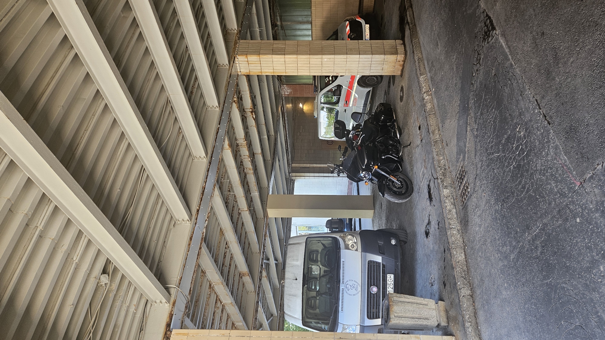 The image depicts a covered parking area, likely in a commercial or industrial zone. Two white vans and a black motorcycle are parked under a corrugated metal roof supported by industrial beams and concrete pillars. One van, on the left, is a delivery van with a license plate that appears to read "125GH". The other van, to the right, is an ambulance or emergency vehicle with red and white markings and a license plate that starts with "BT". The ground is asphalt, with some cracks and oil stains. The overall environment appears utilitarian and somewhat weathered, with visible rust on some of the structural elements. The light suggests it is daytime. The context of Prešov, Slovakia is not evident from the visual information in the image.
