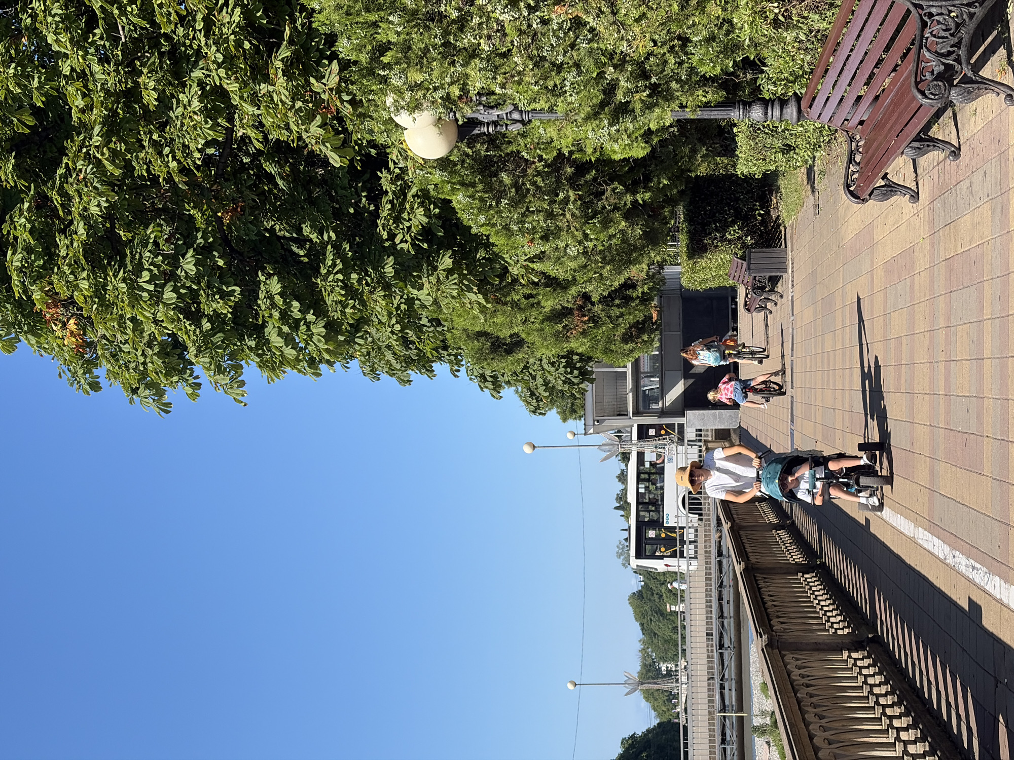 In Sochi, Russia, on a sunny day, two children are riding electric scooters on a paved pathway. The pathway is lined with trees and bushes, and a park bench is visible in the background. A bus is also parked nearby. One child is wearing a straw hat and a white t-shirt, while the other is wearing a pink and white striped shirt and shorts. The children appear to be enjoying their ride, and the overall atmosphere is relaxed and cheerful. The bright blue sky suggests it is daytime.