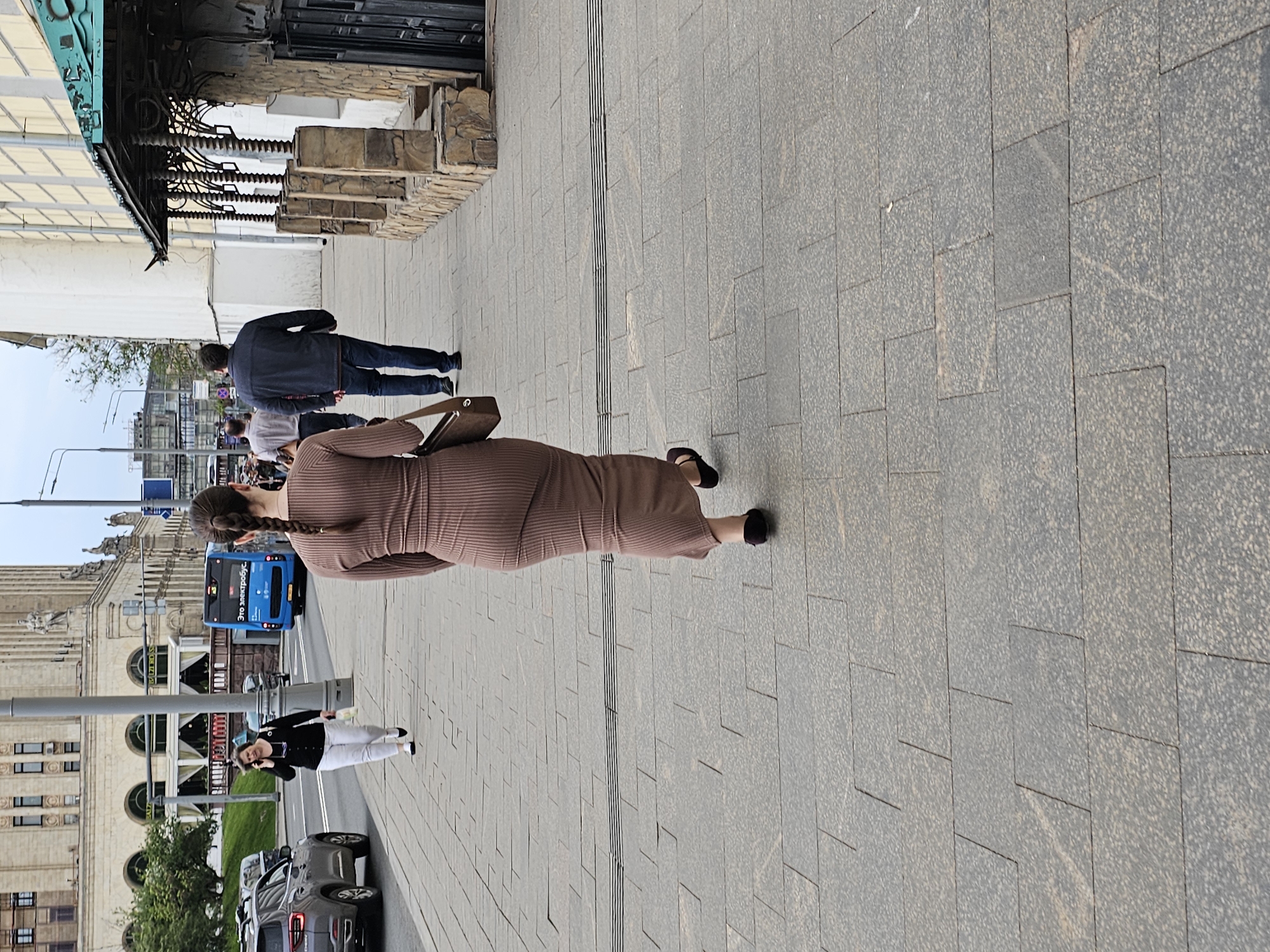 The image depicts a woman in a brown dress walking on a paved sidewalk. She has a brown purse over her shoulder and her hair is braided. Other people are visible in the background, including two men walking together and a woman in a black shirt and white pants talking on her phone. A blue bus is visible further back on the street.
The environment includes buildings, trees, cars, and street lights. The architectural style of the building in the background identifies the location as Moscow, Russia.